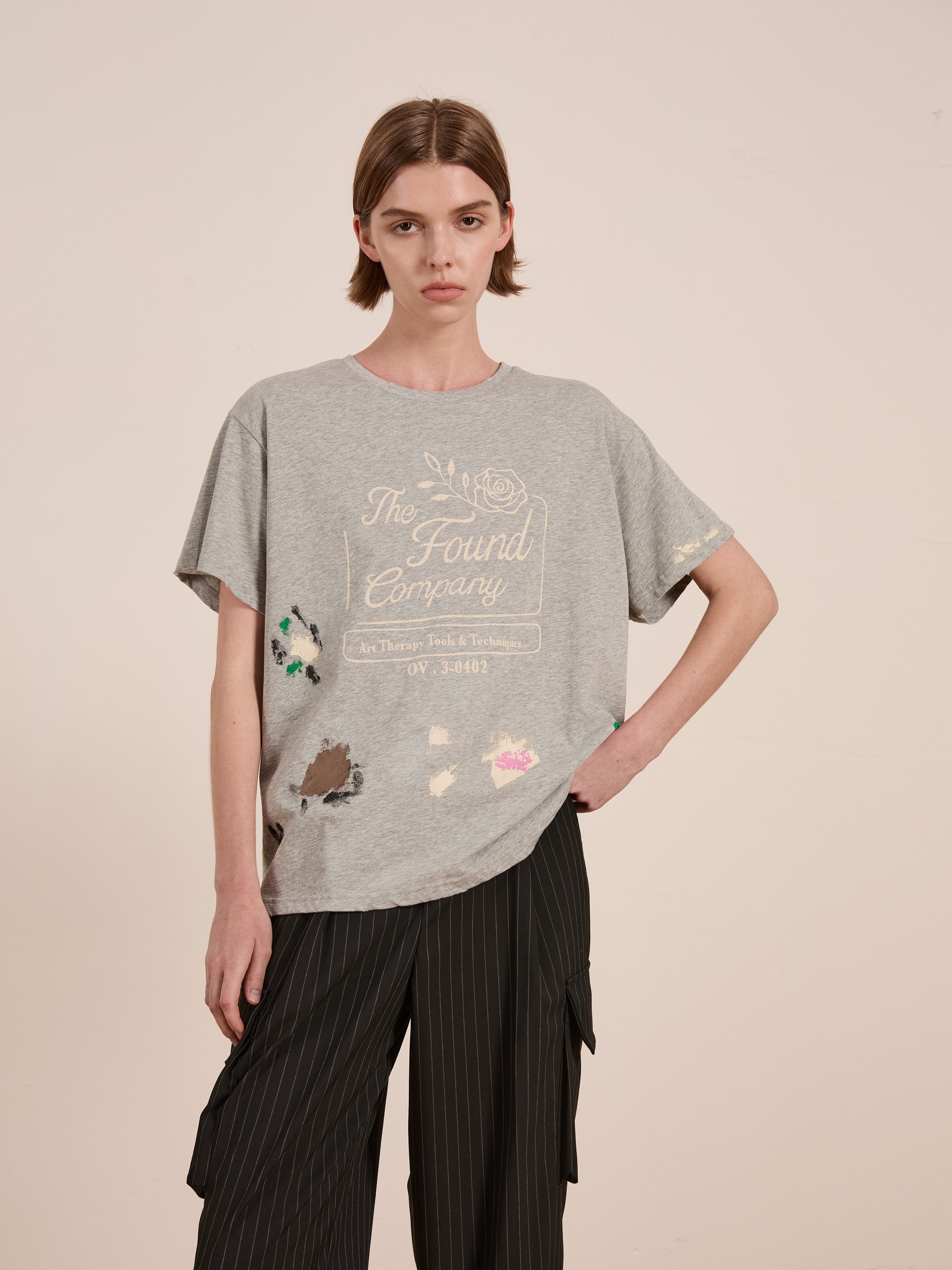 A person with short brown hair stands against a plain backdrop, dressed in a gray Painters Company Tee by FOUND, showcasing vintage-inspired graphics, and black pinstripe pants.