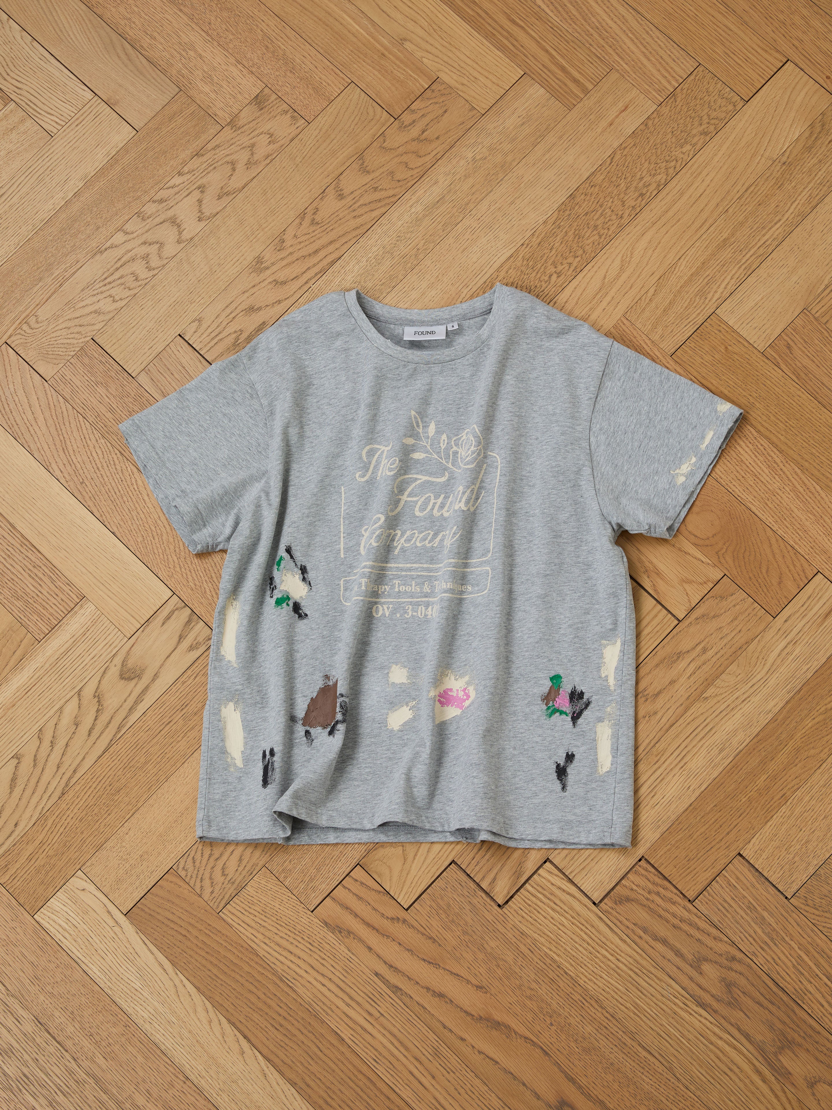 The Painters Company Tee by FOUND, a gray unisex T-shirt with colorful paint splatters and white text, exudes vintage-inspired charm while laid on a wooden parquet floor.