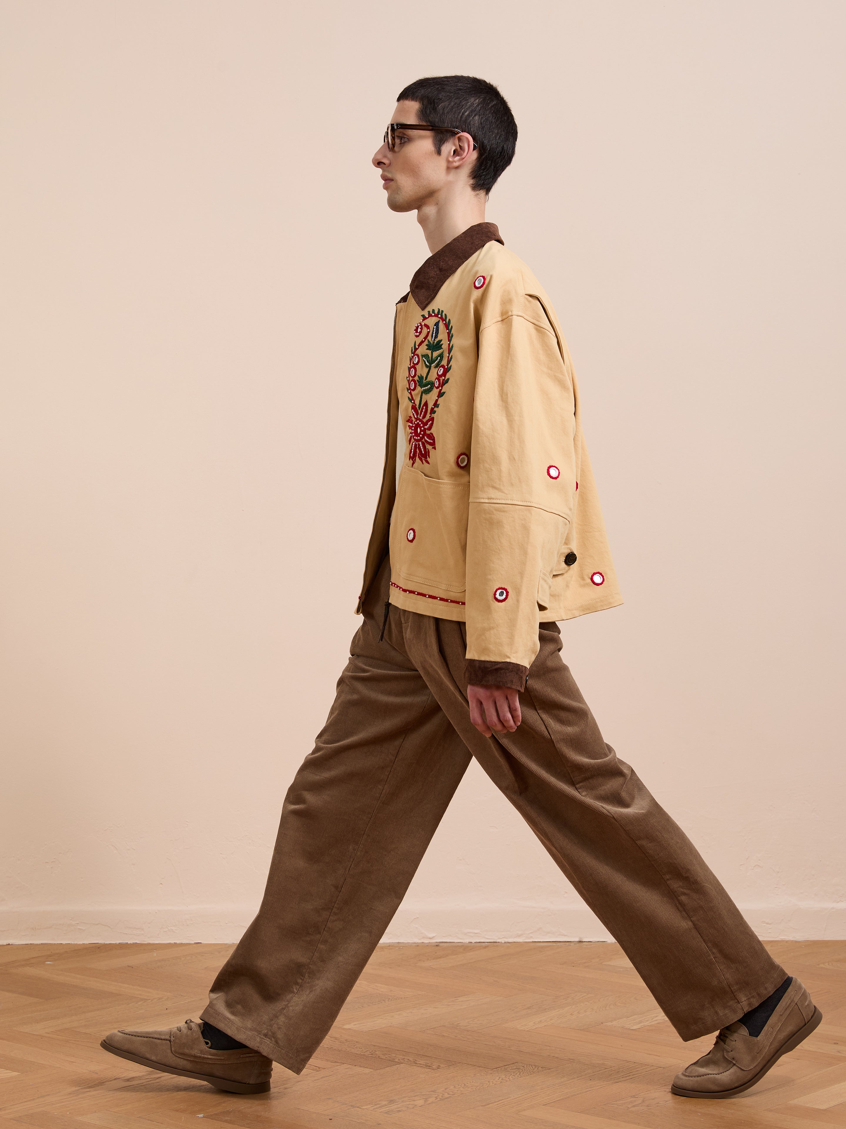 A person in glasses walks indoors wearing the Found Paisley Motif Hand Embroidered Work Jacket, paired with brown corduroy pants and tan shoes, highlighting artisan craftsmanship against a plain wall and wood floor.