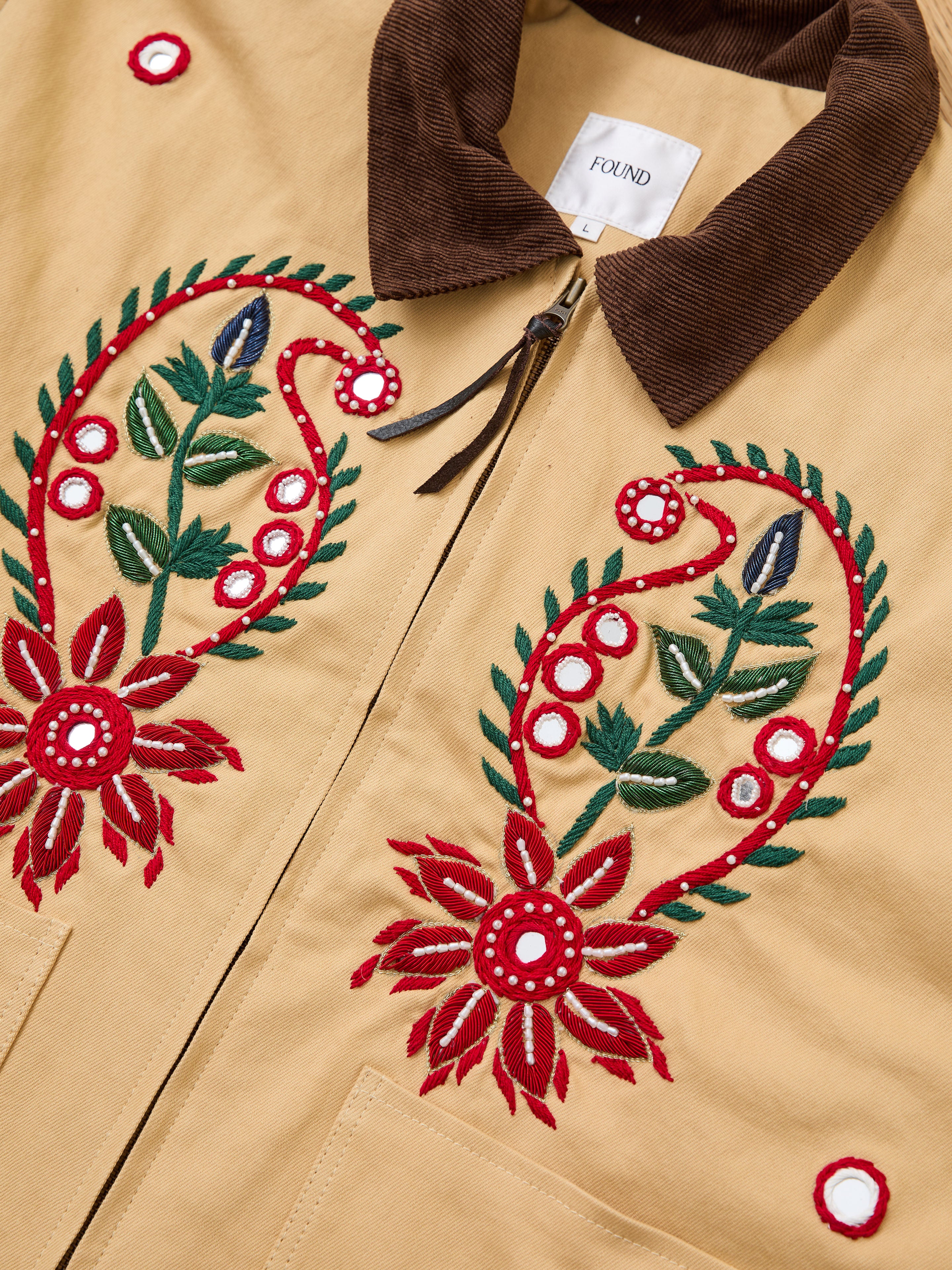The Found Paisley Motif Hand Embroidered Work Jacket is tan with a brown corduroy collar, artisan red, green, and white paisley floral embroidery on the front, a visible zipper, and the Found label inside the collar.