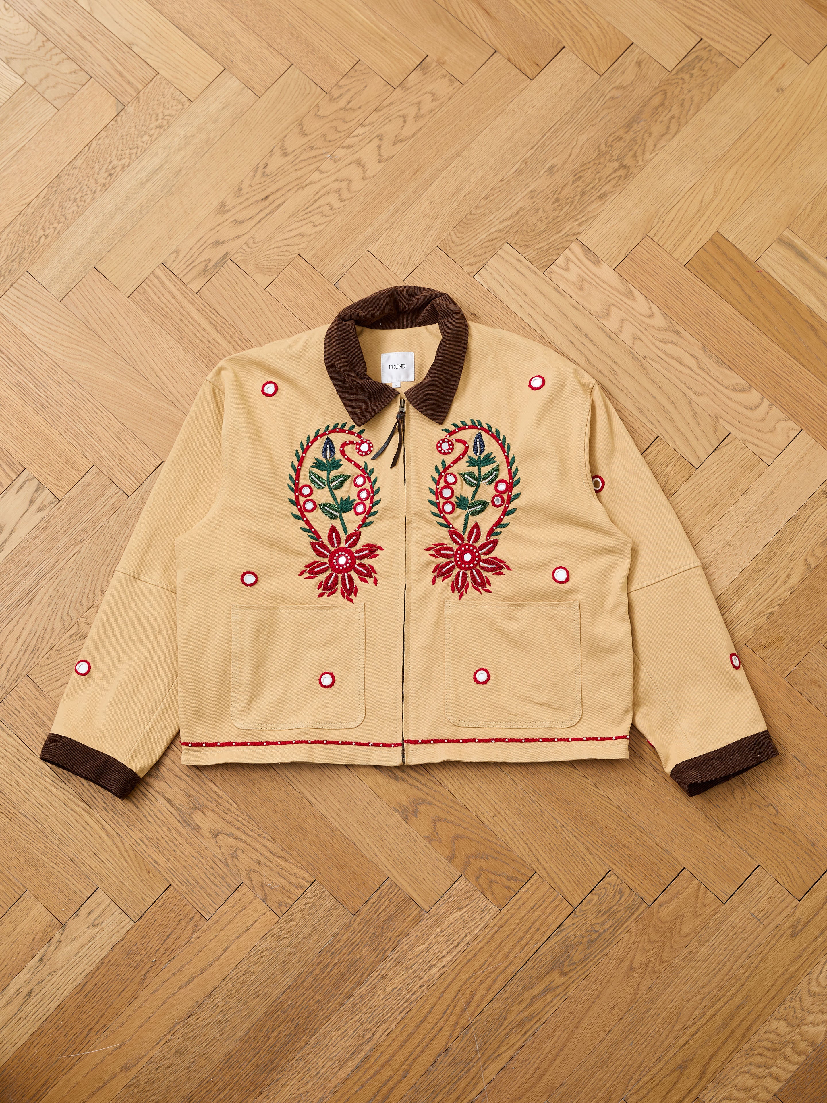 The Found Paisley Motif Hand Embroidered Work Jacket features tan fabric, a brown collar and cuffs, paisley floral embroidery, and red circular accents. It’s displayed flat on a parquet floor, showcasing its artisan craftsmanship.