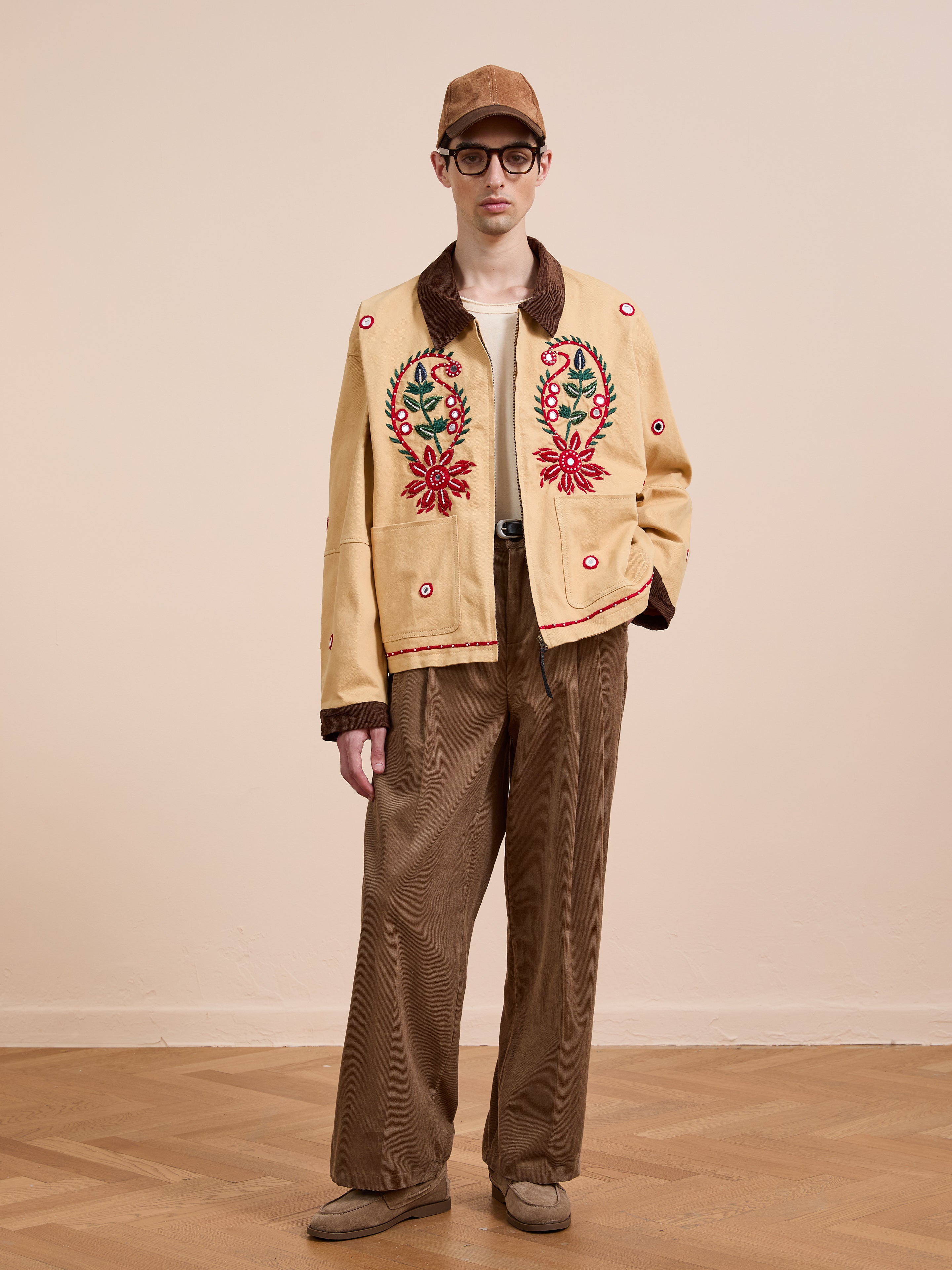 A person wearing the Found Paisley Motif Hand Embroidered Work Jacket stands on a wooden floor against a plain beige wall, styled with brown corduroy pants, a beige shirt, brown cap, and glasses.