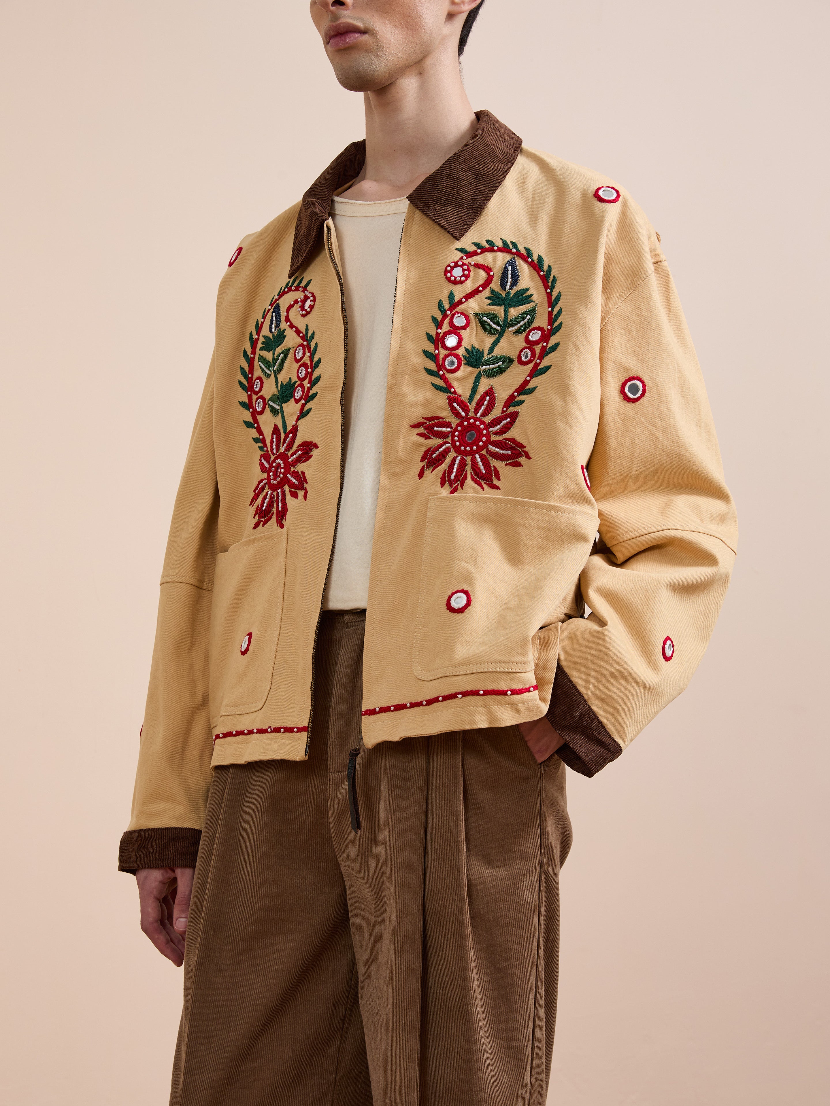 A person wears Found's Paisley Motif Hand Embroidered Work Jacket—beige with red and green paisley floral embroidery, brown collar and cuffs—paired with brown trousers, standing against a plain background.