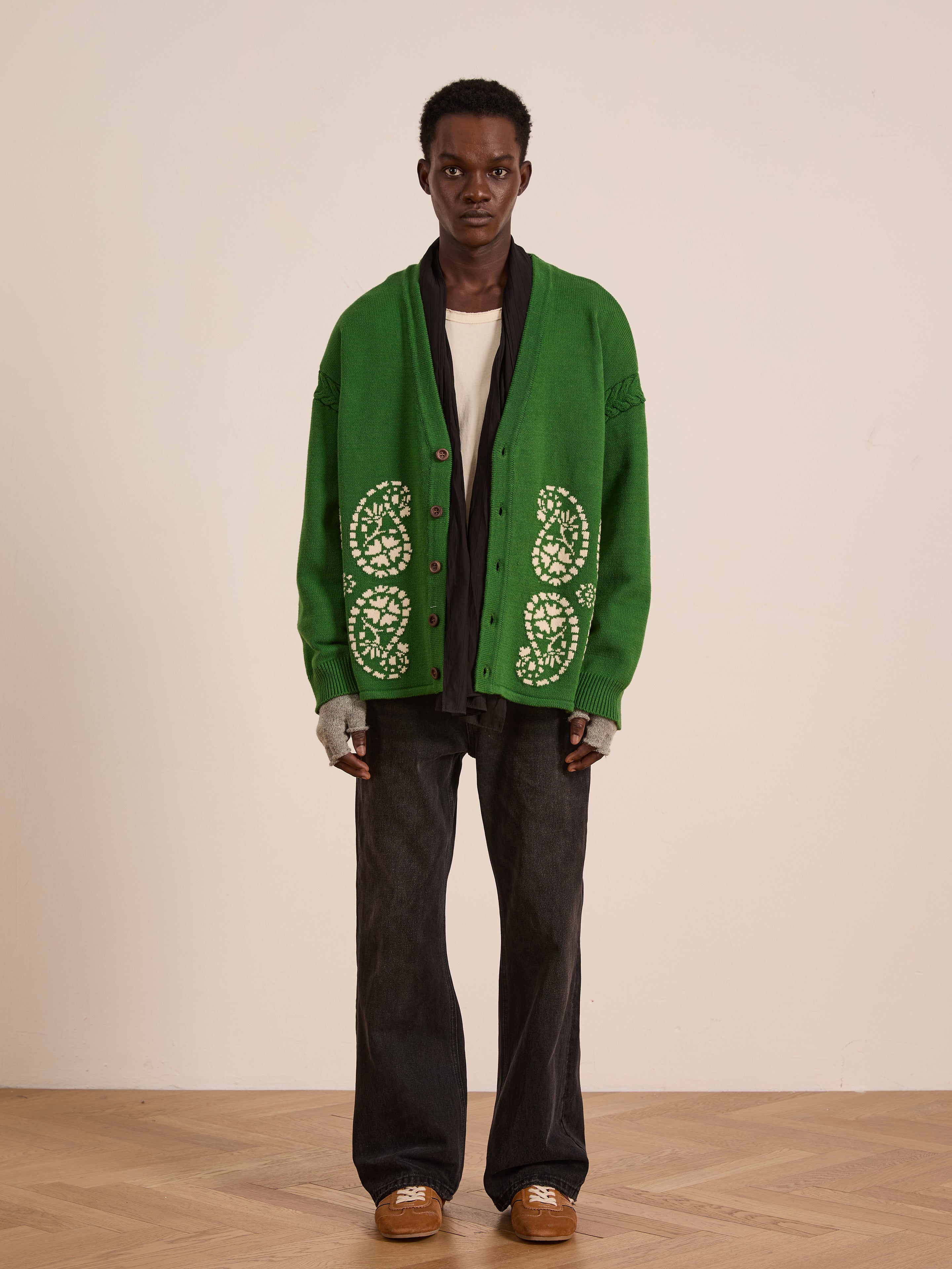 A person stands indoors against a plain background, facing forward, wearing the FOUND Paisley Knit Cardigan in green with white patterns, paired with a white t-shirt, black pants, brown shoes, and fingerless gloves.