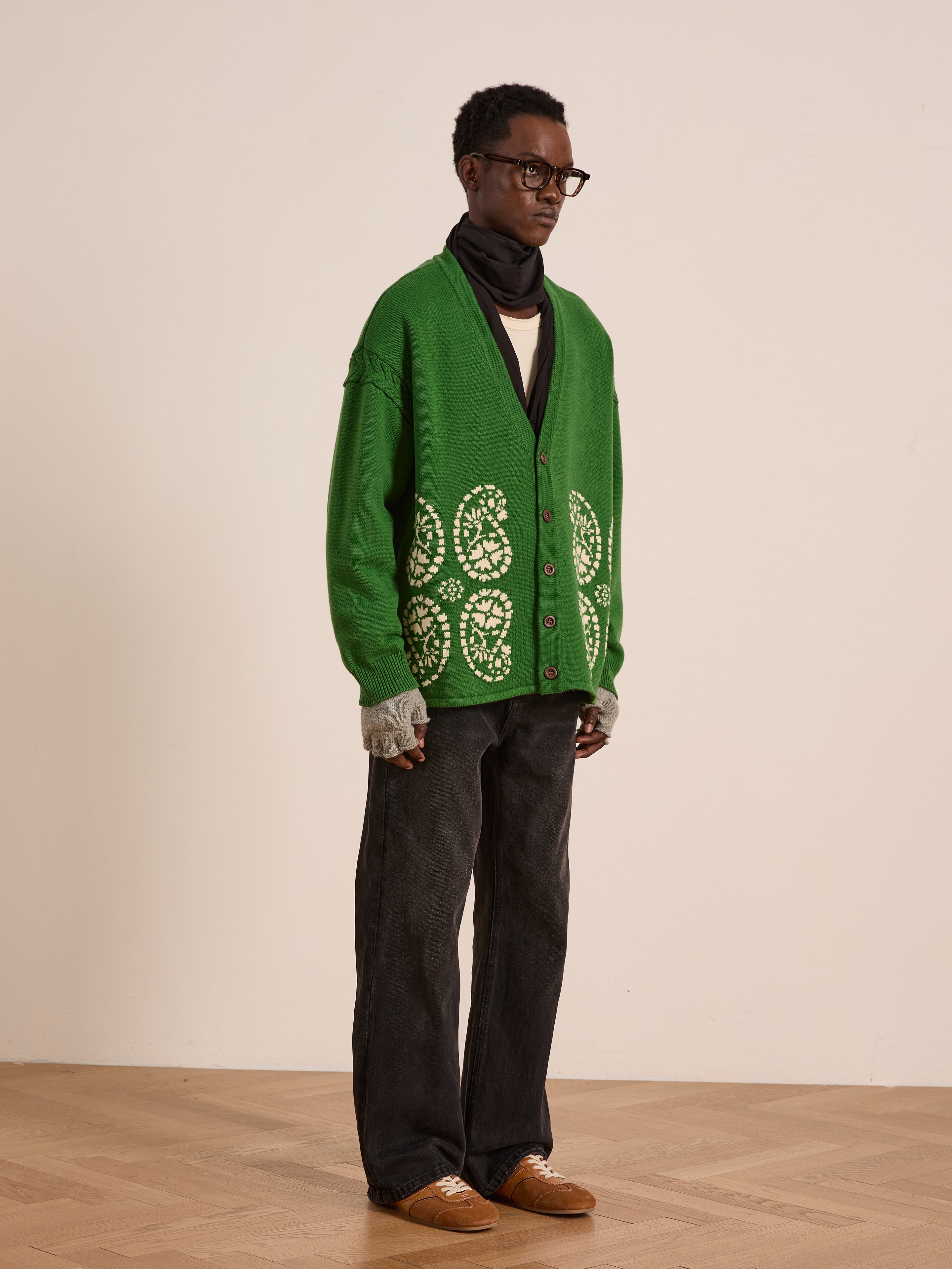 A person wearing FOUND’s green Paisley Knit Cardigan, dark jeans, gray gloves, brown shoes, glasses, and a black scarf stands indoors on a wooden floor against a plain wall.