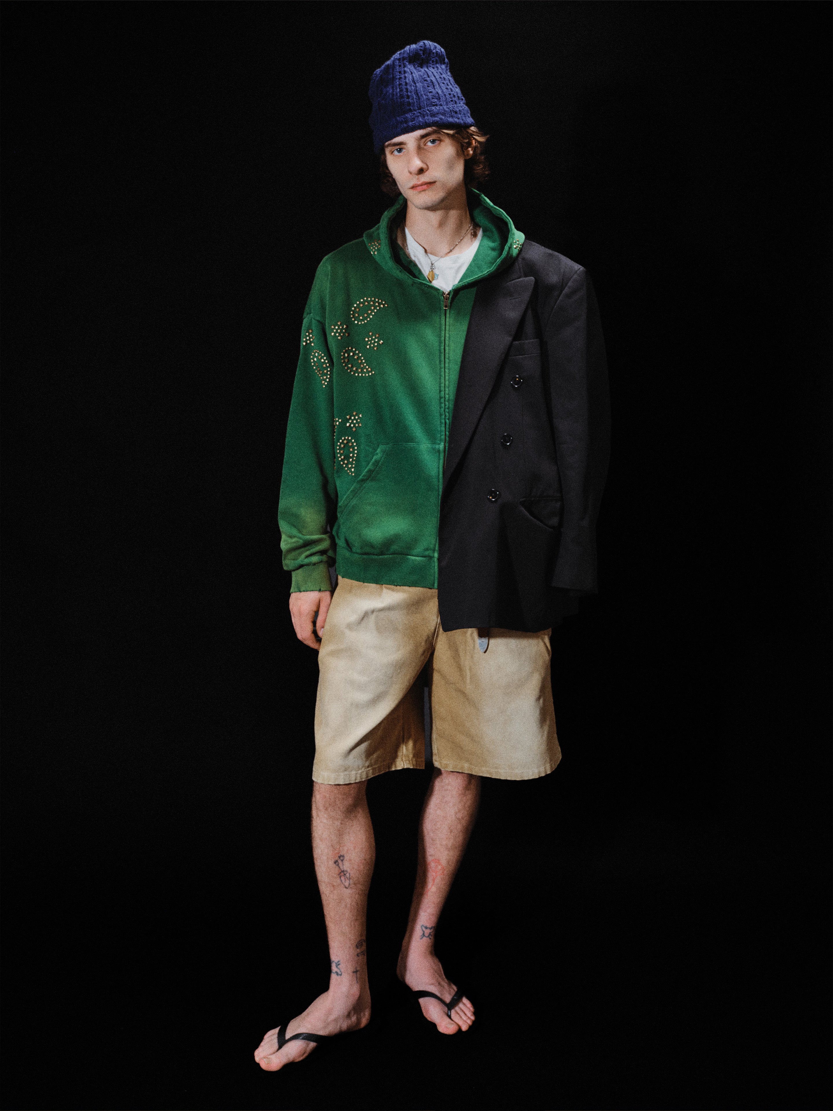 A person wearing a blue beanie, the FOUND Paisley Studded Zip Up Hoodie in green, a black blazer, beige shorts, and flip-flops stands against a black background.