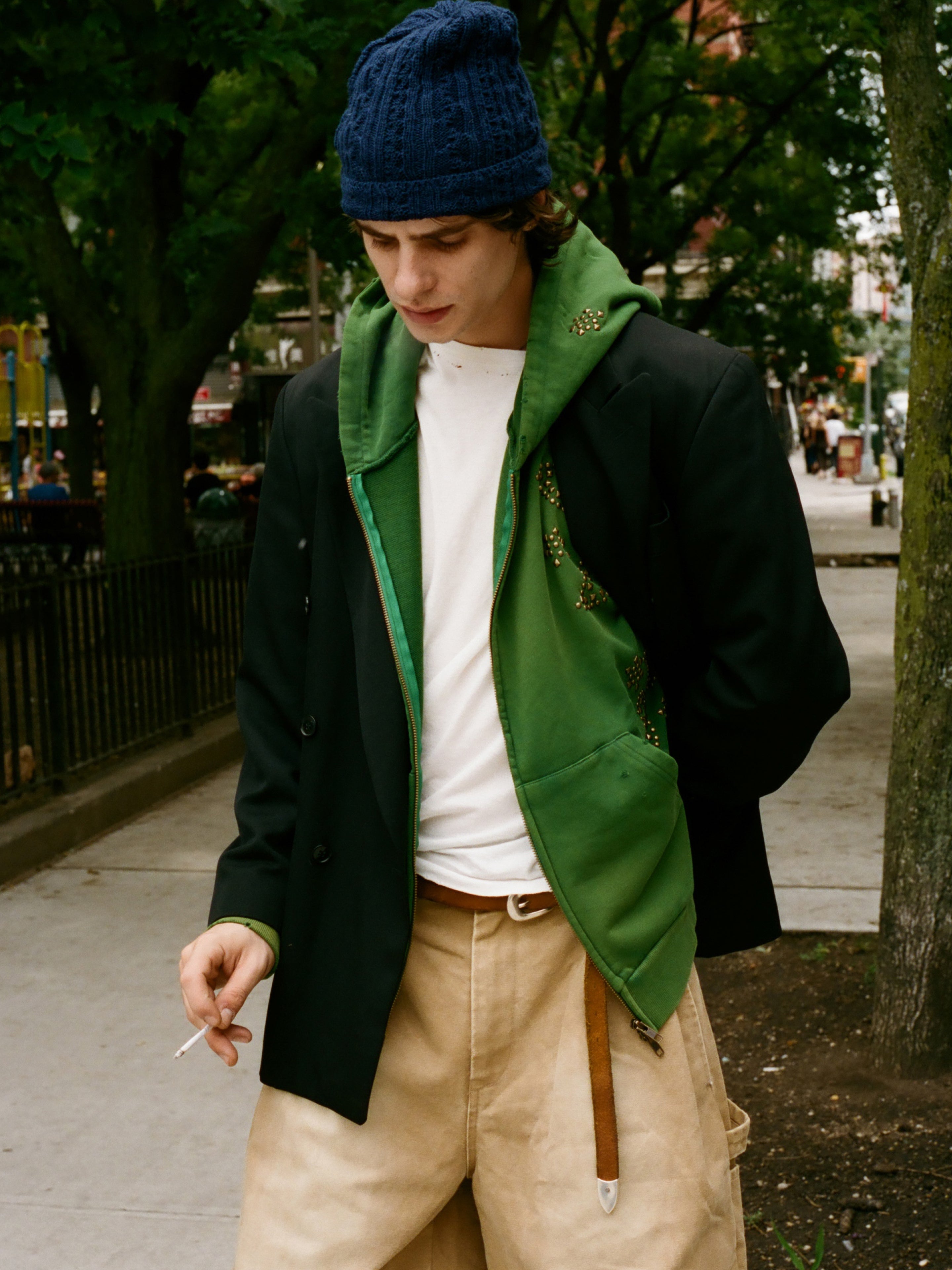 A person wearing a blue knit hat, a FOUND Paisley Studded Zip Up Hoodie in green layered under a black coat, a white shirt, and beige pants stands outdoors holding a cigarette.