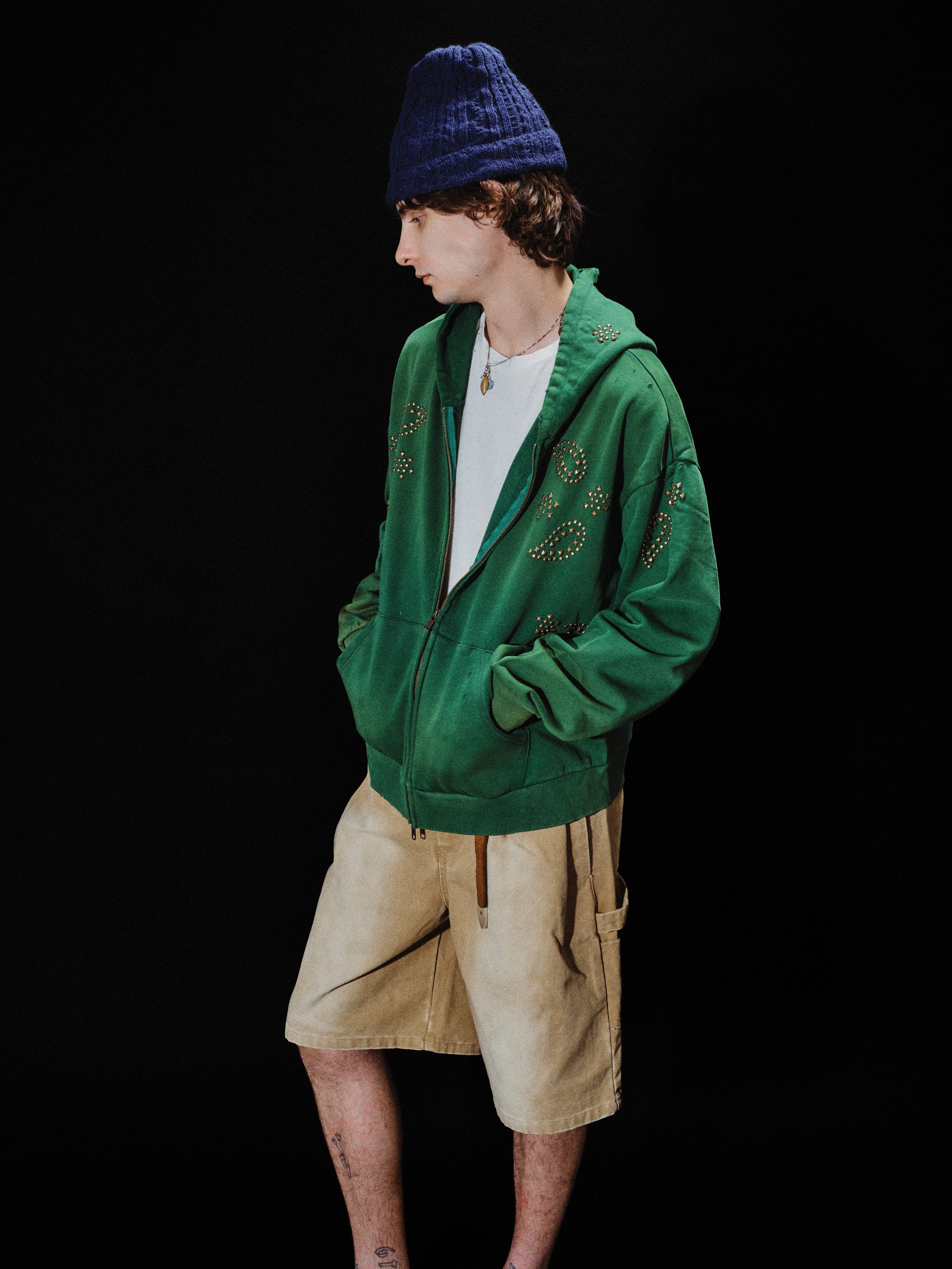 A person wearing the FOUND Paisley Studded Zip Up Hoodie in vintage green, along with a white t-shirt, beige shorts, and a blue knit hat, stands with hands in pockets against a black background.