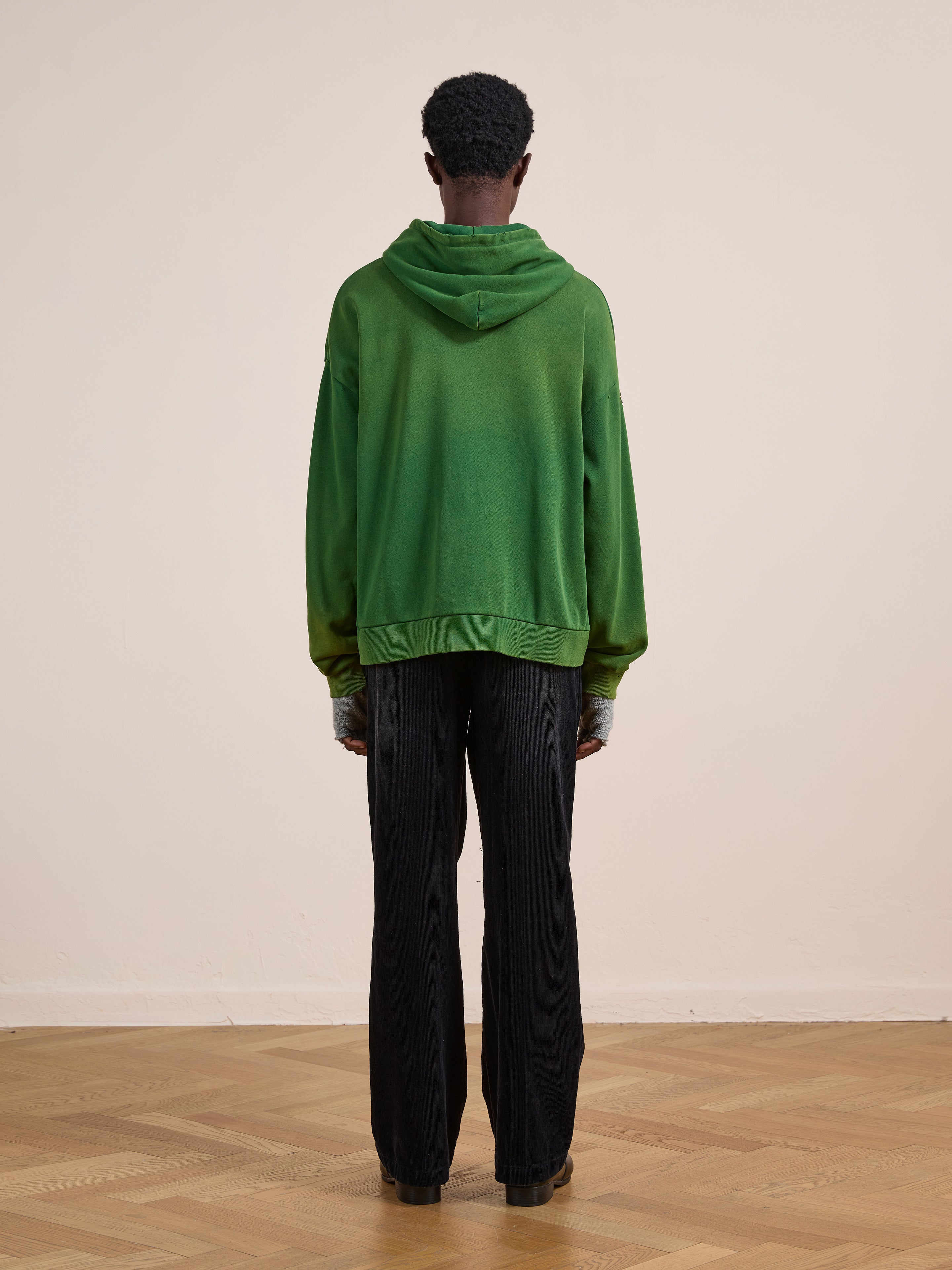 A person stands indoors facing away, wearing the FOUND Paisley Studded Zip Up Hoodie in a unisex relaxed fit, paired with black pants and brown shoes on a wooden floor against a plain, light-colored wall.