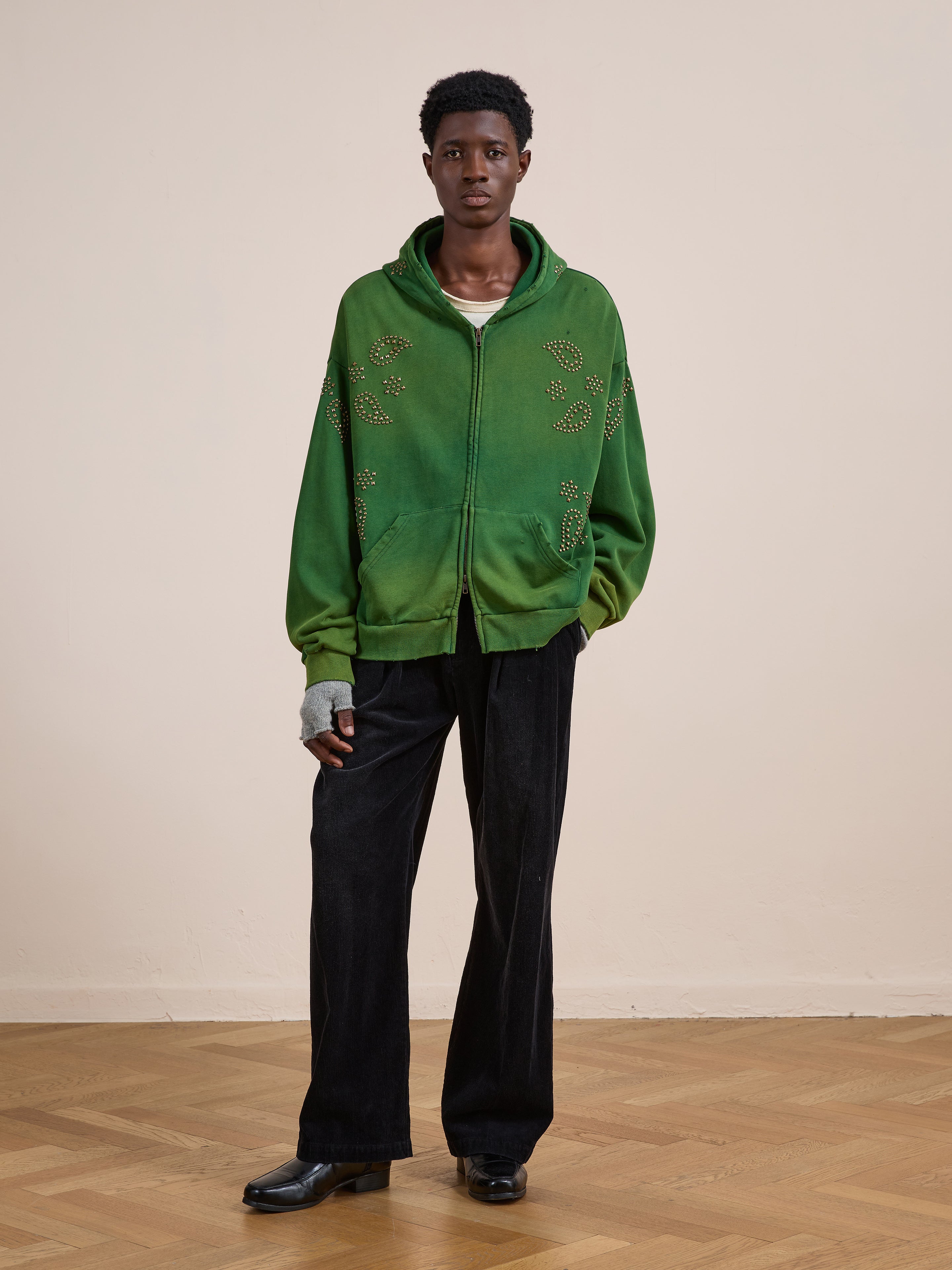 A person indoors sports the FOUND Paisley Studded Zip Up Hoodie—green with decorative patterns—paired with black wide-leg pants, black shoes, and a gray fingerless glove for a cool, vintage look.