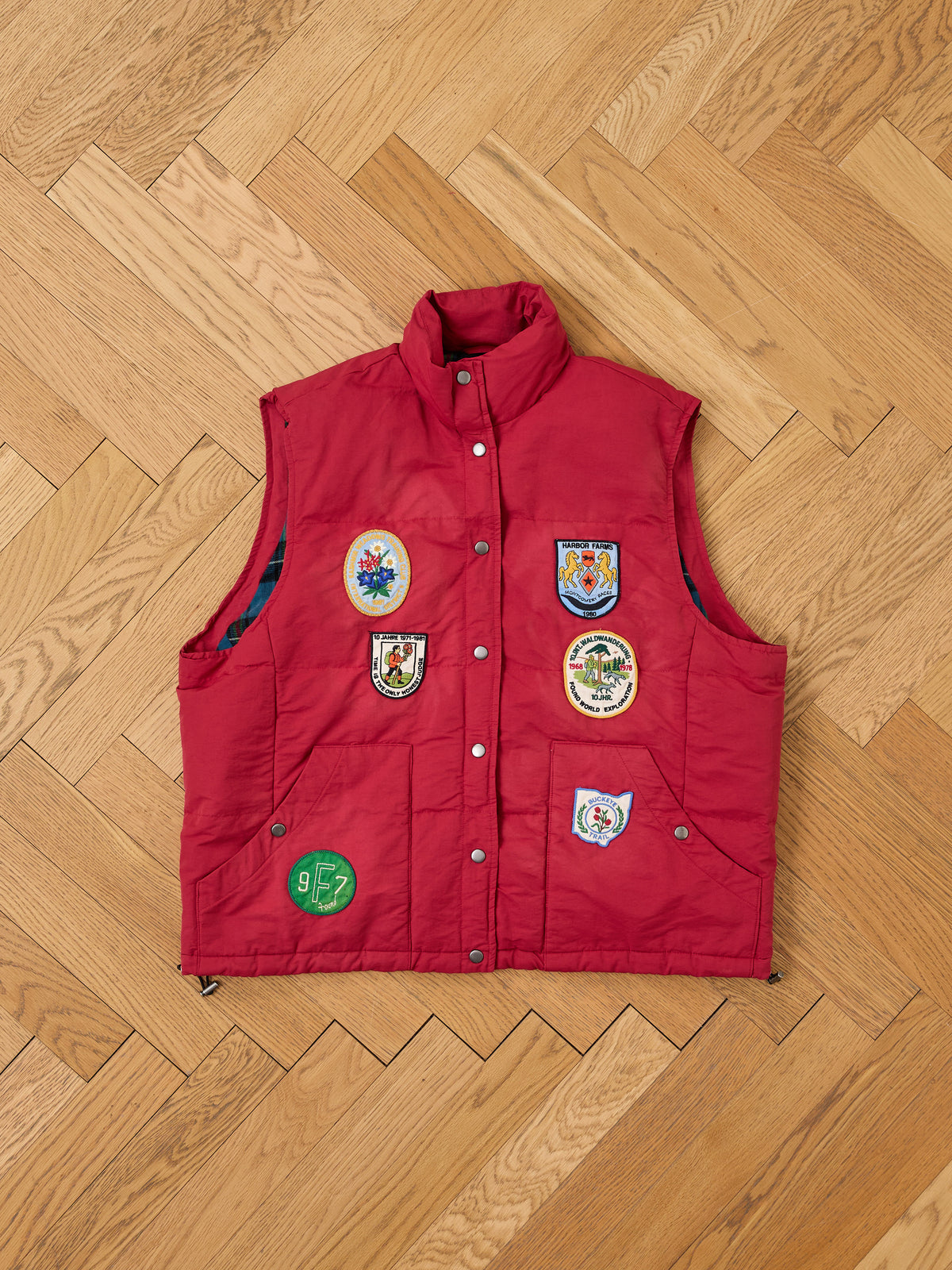 The FOUND Patch Down Vest in red features colorful embroidered patches, showcased on a light wood herringbone floor.