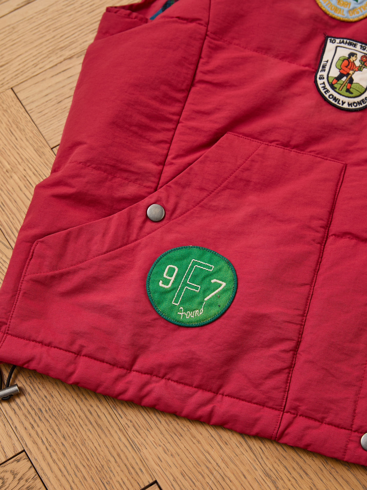 FOUND Patch Down Vest featuring bold red color and embroidered patches, including a green “9 F 7” circle, shown on a wood floor. Great outdoor style.