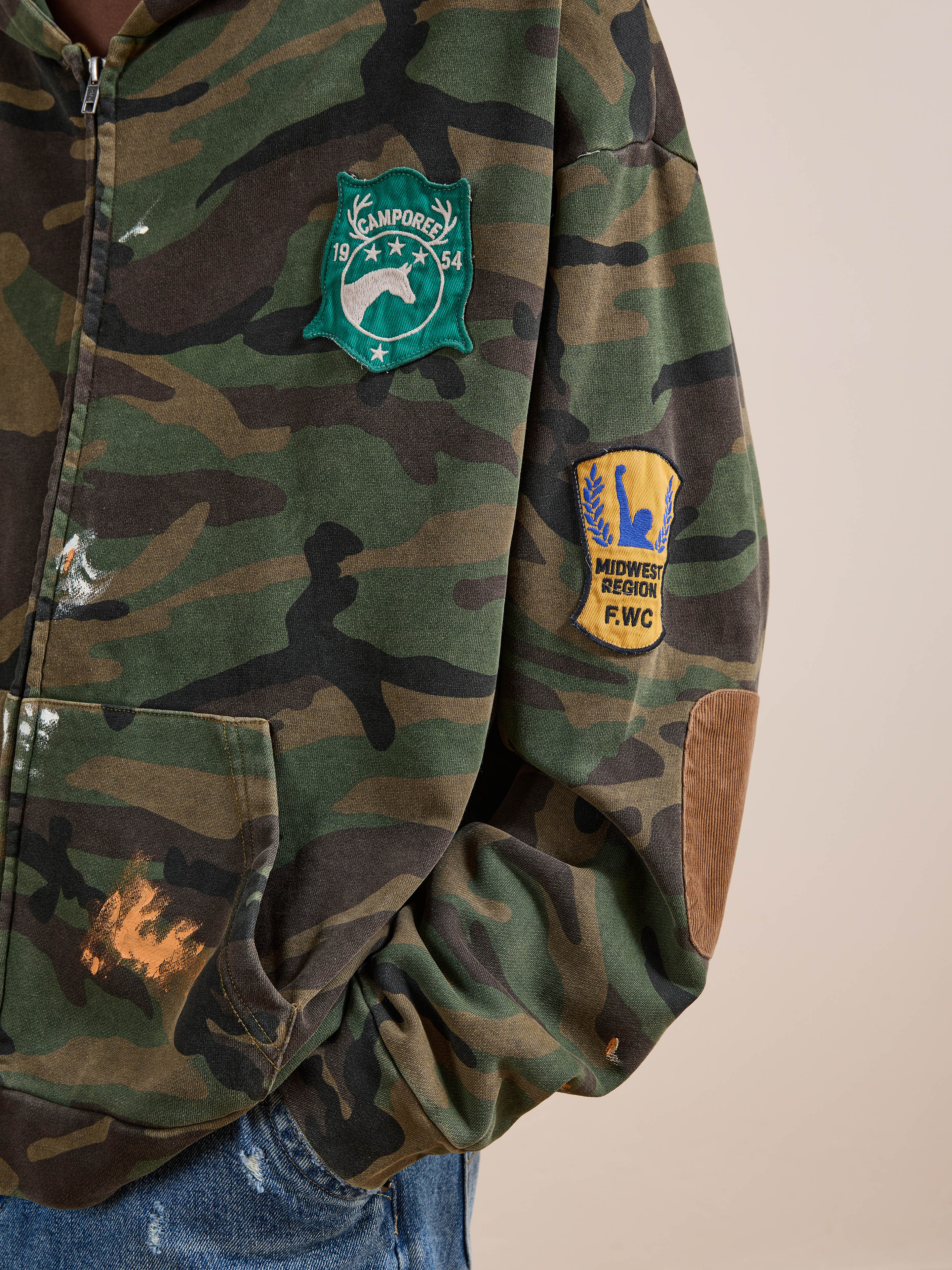 Wearing the FOUND Patch Camo Zip Up Hoodie, a relaxed fit unisex style featuring a green "Camporee 1954" patch on the chest and a yellow "Midwest Region F.W.C." patch on the sleeve.