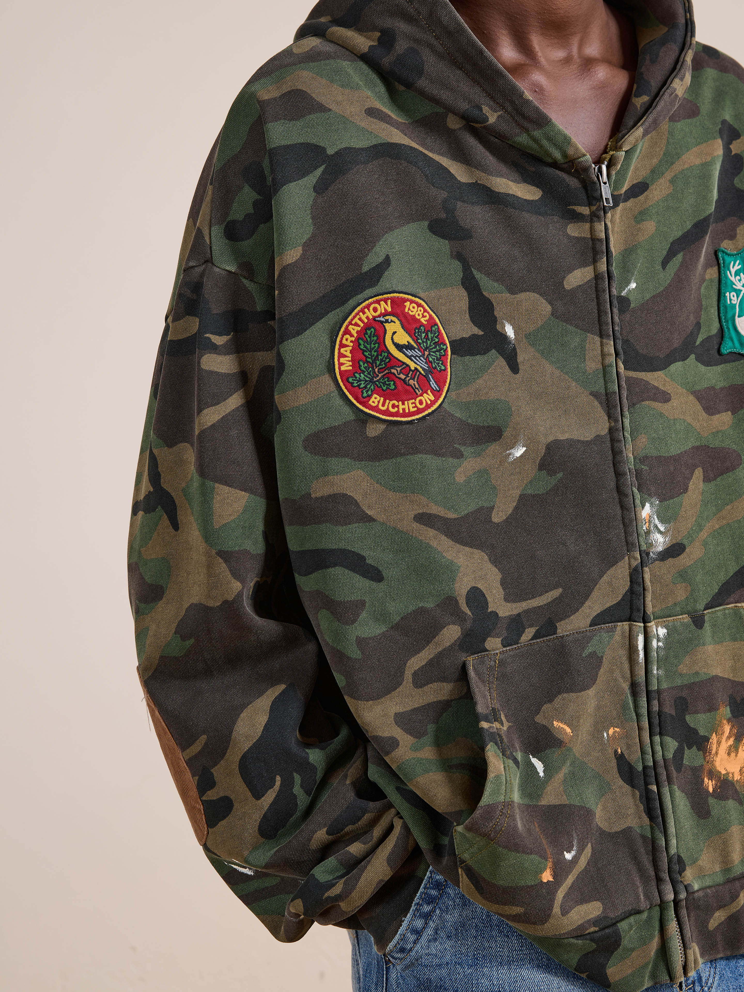 A person wears the FOUND Patch Camo Zip Up Hoodie, featuring a "Marathon Bucheon 1982" patch and a green patch, styled in a relaxed fit with blue jeans.