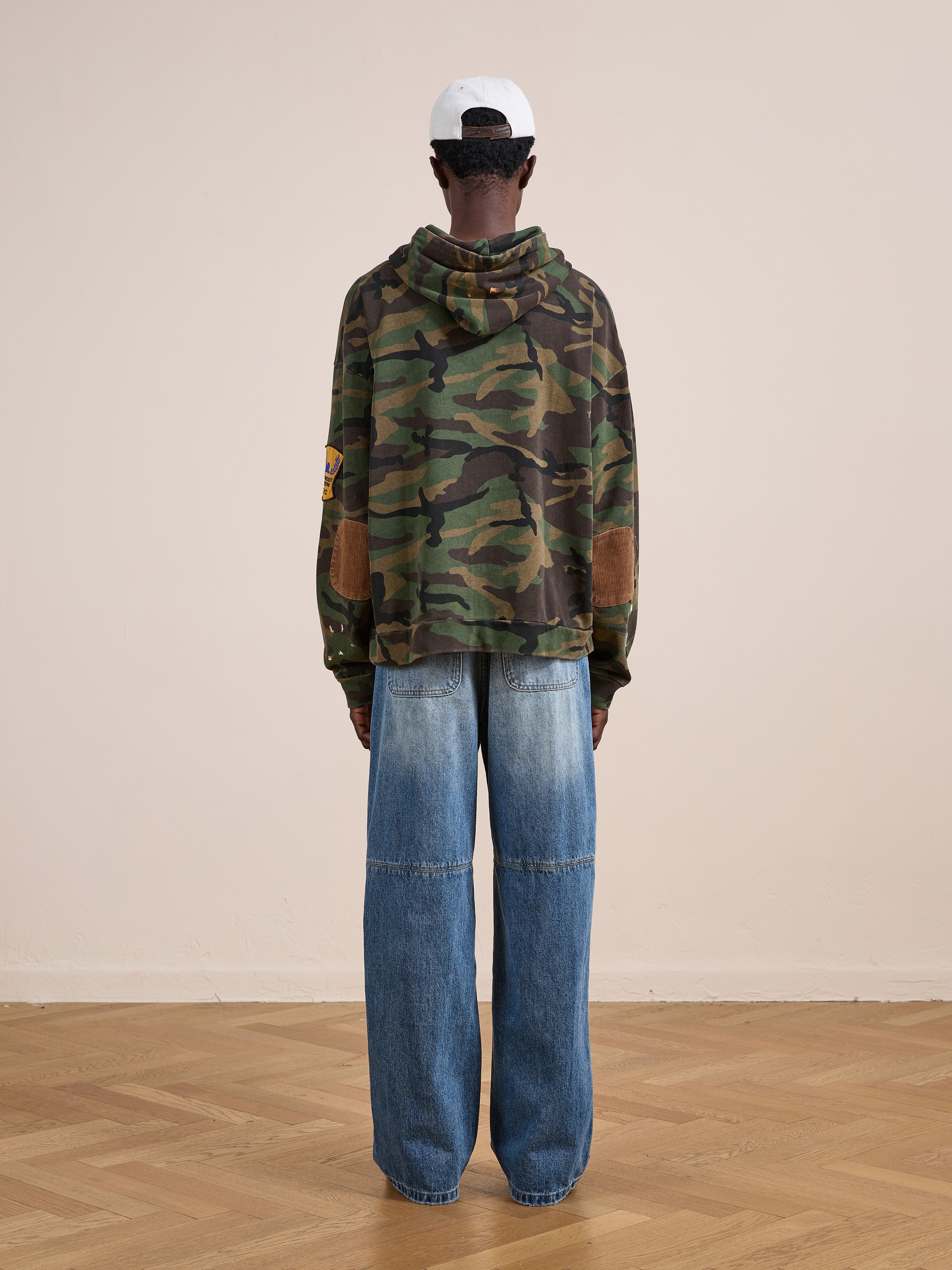 A person indoors faces away, wearing the FOUND Patch Camo Zip Up Hoodie with loose blue jeans and a white baseball cap. The wooden floor and light wall enhance the hoodie’s relaxed, unisex style.