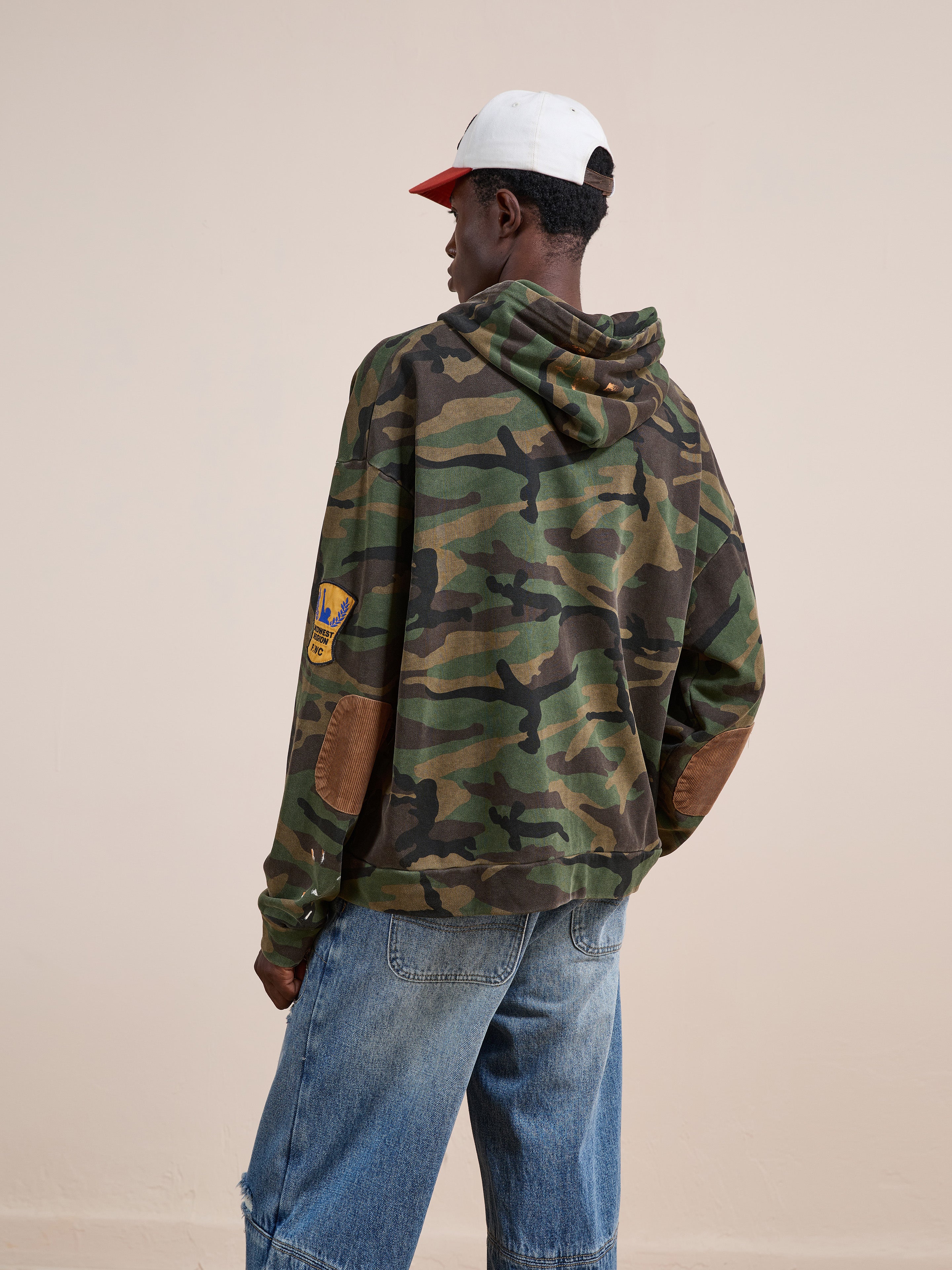 A person wearing the FOUND Patch Camo Zip Up Hoodie, blue jeans, and a white and red cap stands against a plain beige background, facing away from the camera.