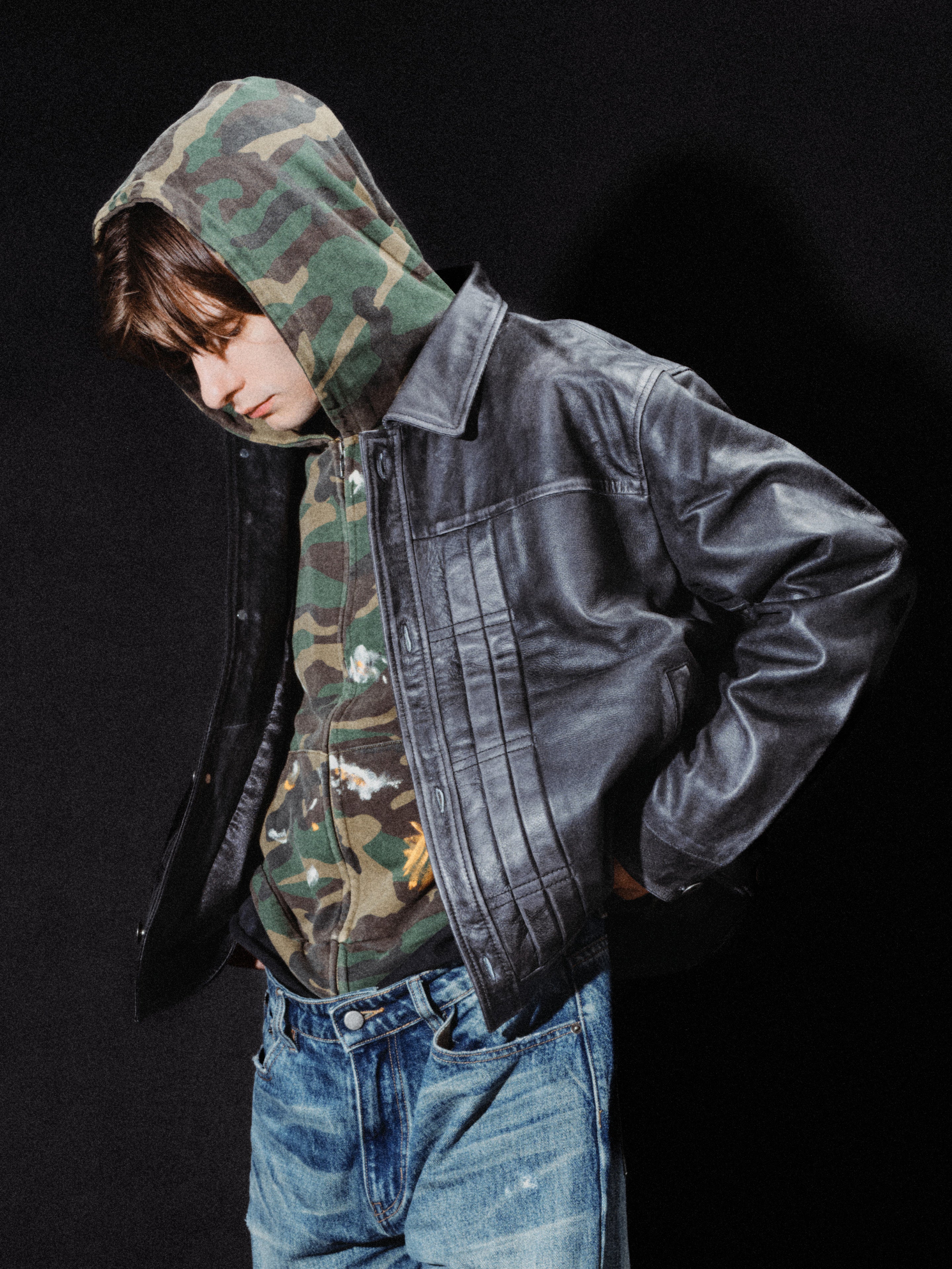 Wearing the FOUND Patch Camo Zip Up Hoodie in a unisex relaxed fit layered under a black leather jacket and paired with blue jeans, the person stands against a black background, looking downward.
