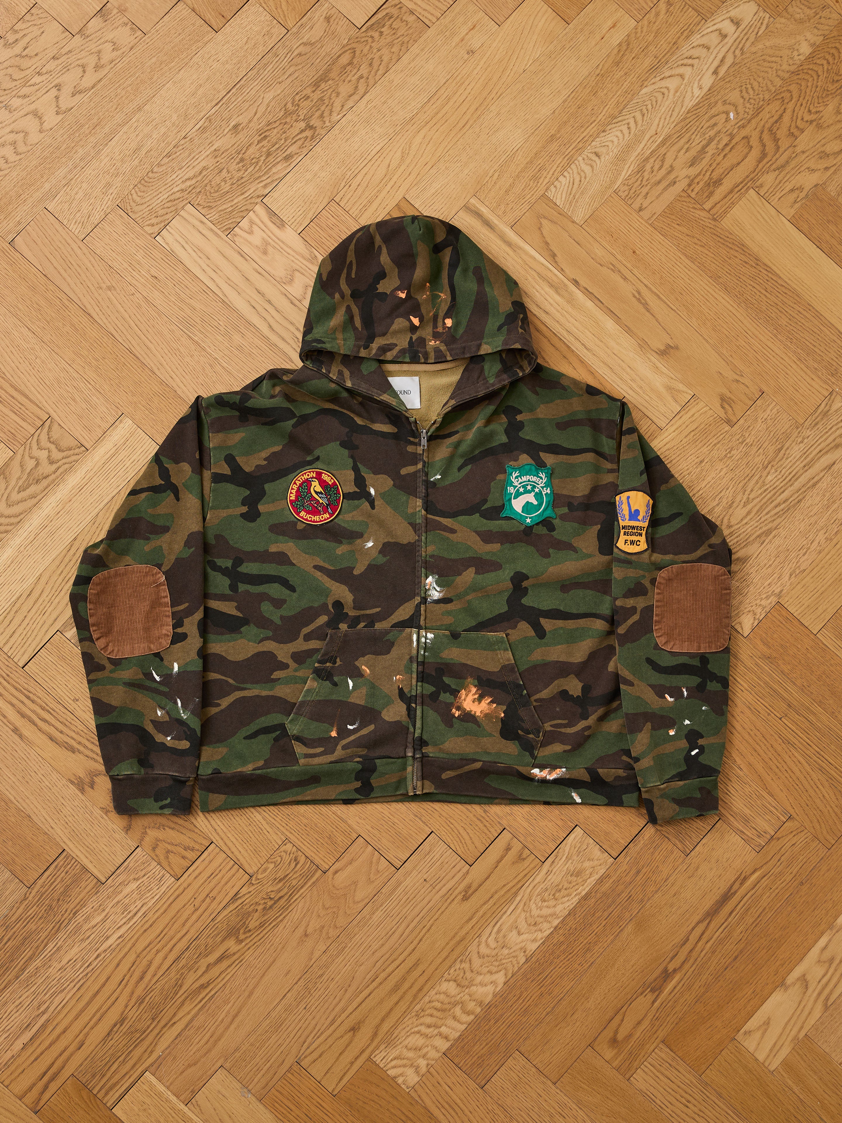 The FOUND Patch Camo Zip Up Hoodie, featuring a relaxed fit, hood, front zipper, elbow patches, and three sewn patches on the chest and sleeve, is displayed flat on a wooden herringbone floor.