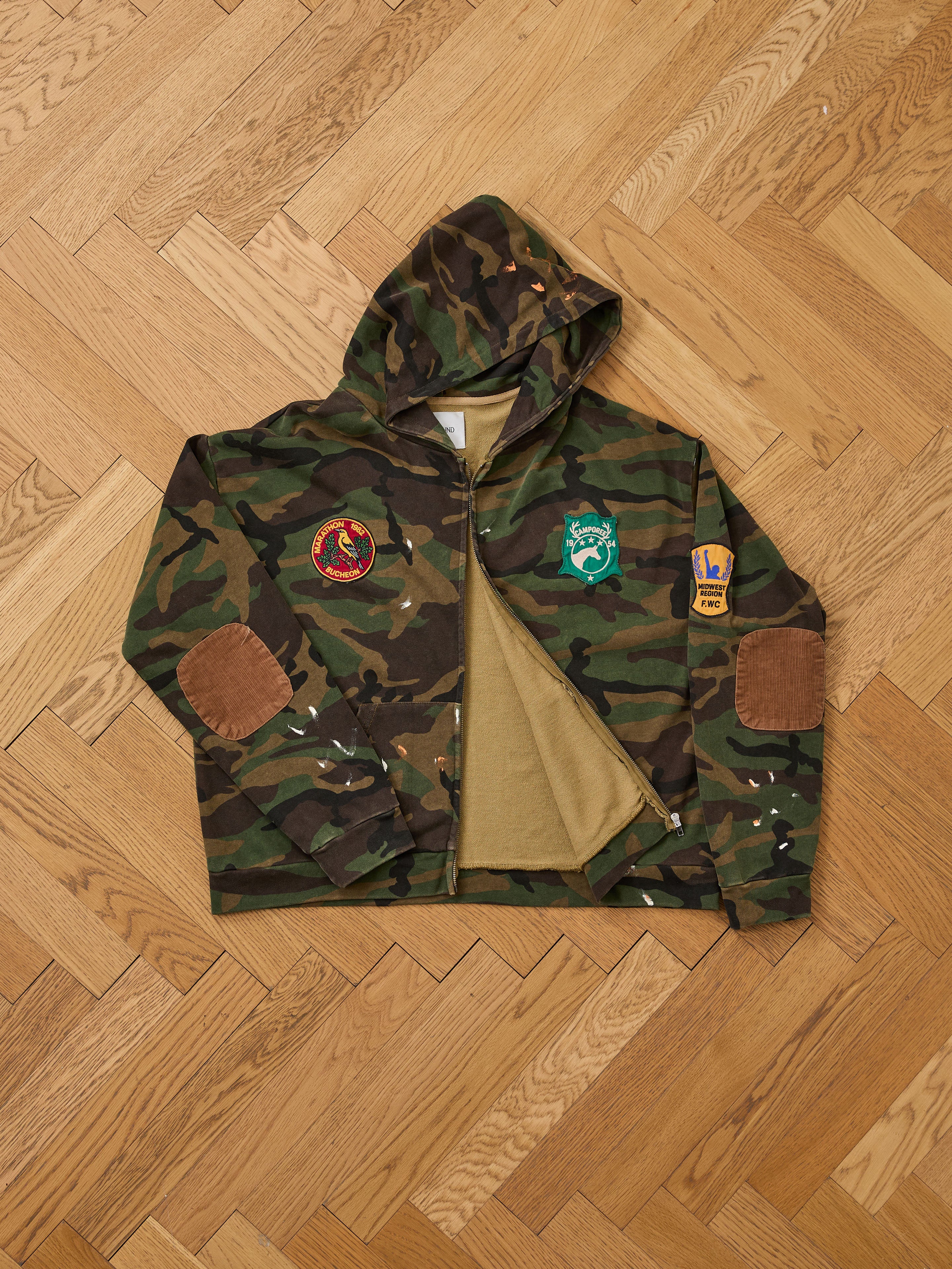 The FOUND Patch Camo Zip Up Hoodie features a relaxed fit with elbow and three embroidered patches, shown on a wooden herringbone floor.