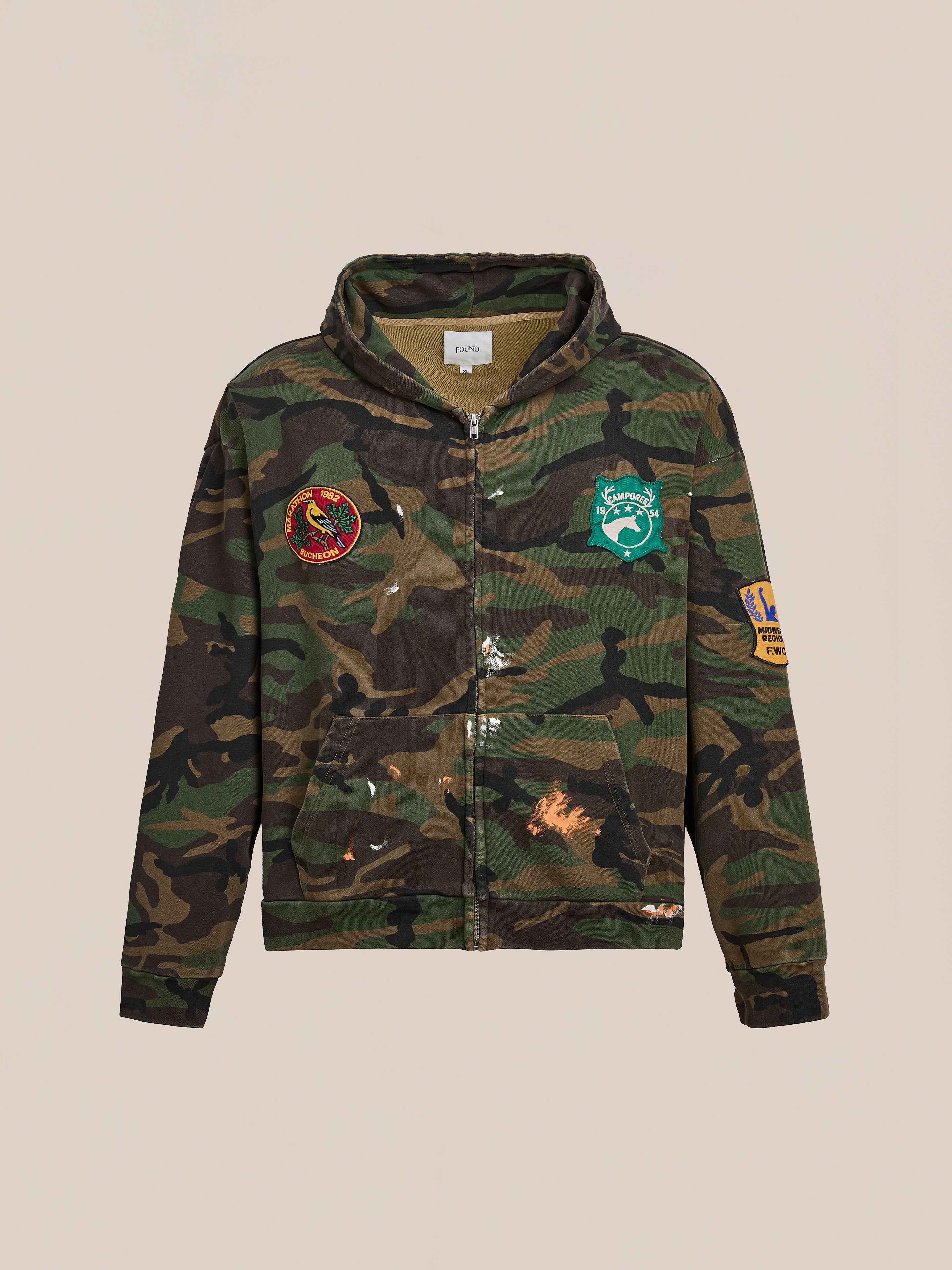 The FOUND Patch Camo Zip Up Hoodie features a relaxed fit with colorful patches and paint splatter details, shown against a plain light background.