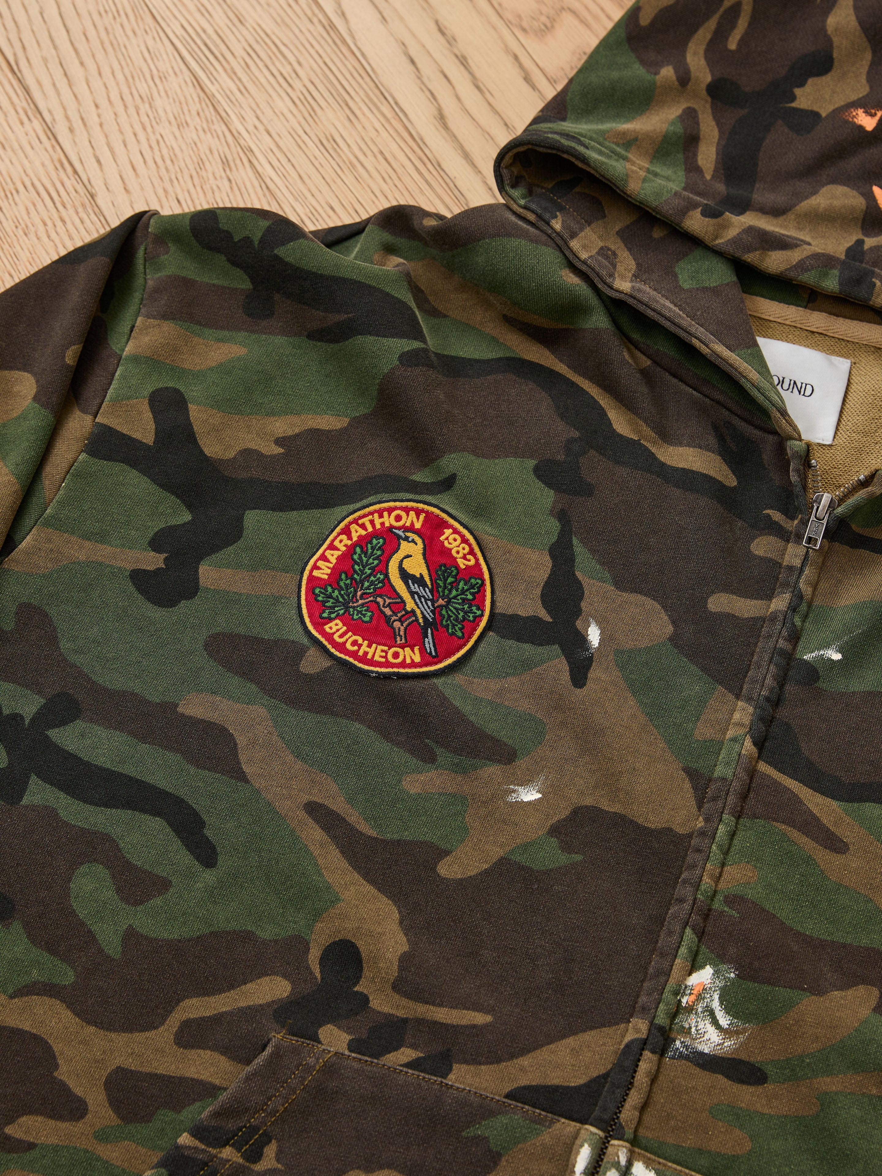 The FOUND Patch Camo Zip Up Hoodie is a unisex, relaxed-fit hoodie with a front zipper and a circular "MARATHON 1982 BUCHEON" patch showing a parrot and leaves. It’s displayed flat on a wooden surface.