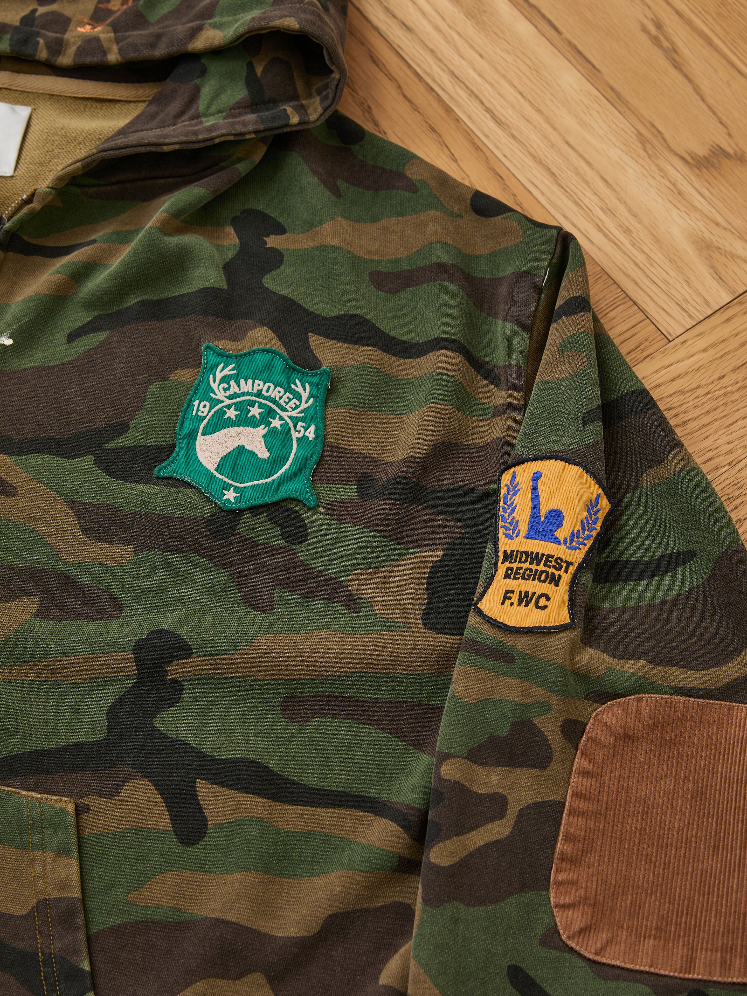 The FOUND Patch Camo Zip Up Hoodie features a relaxed fit, a green Camporee 1954 chest patch, and a yellow Midwest Region F.W.C. sleeve patch, shown laid on a wooden floor.