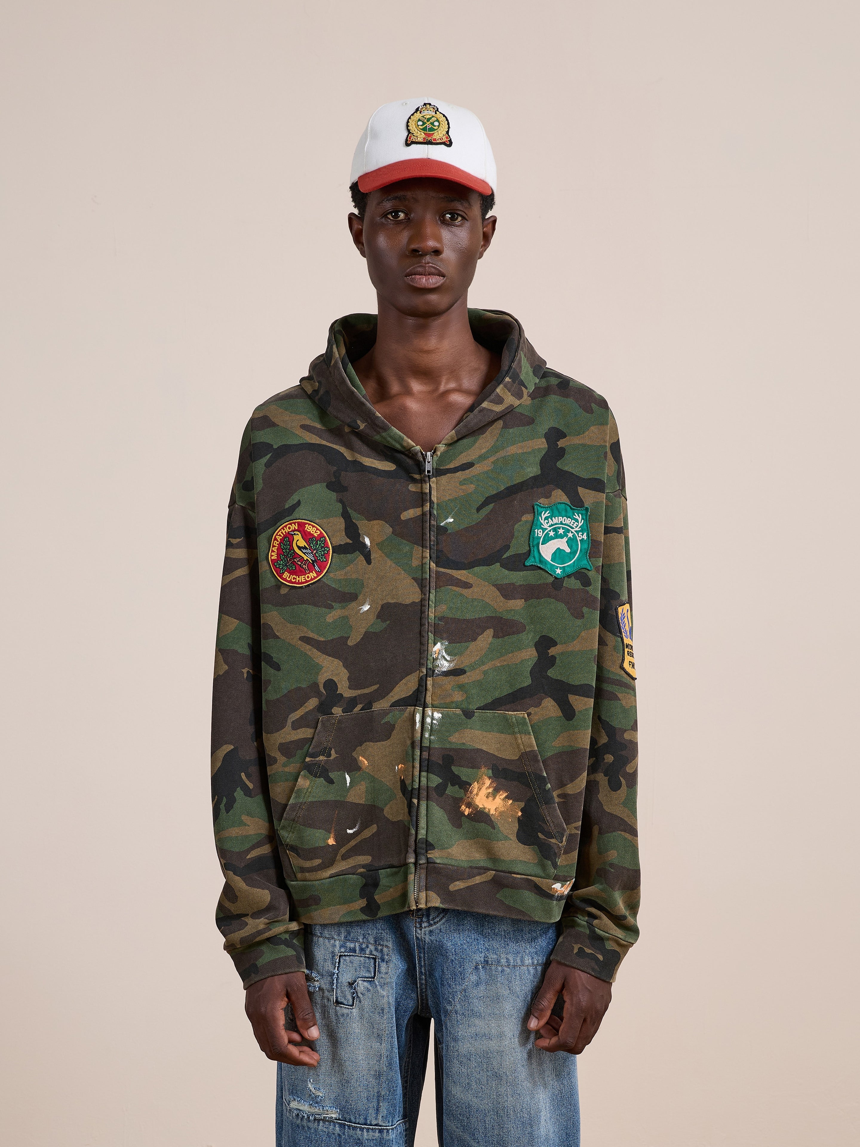 A person wearing the FOUND Patch Camo Zip Up Hoodie, styled with blue jeans and a white baseball cap, stands against a plain background.