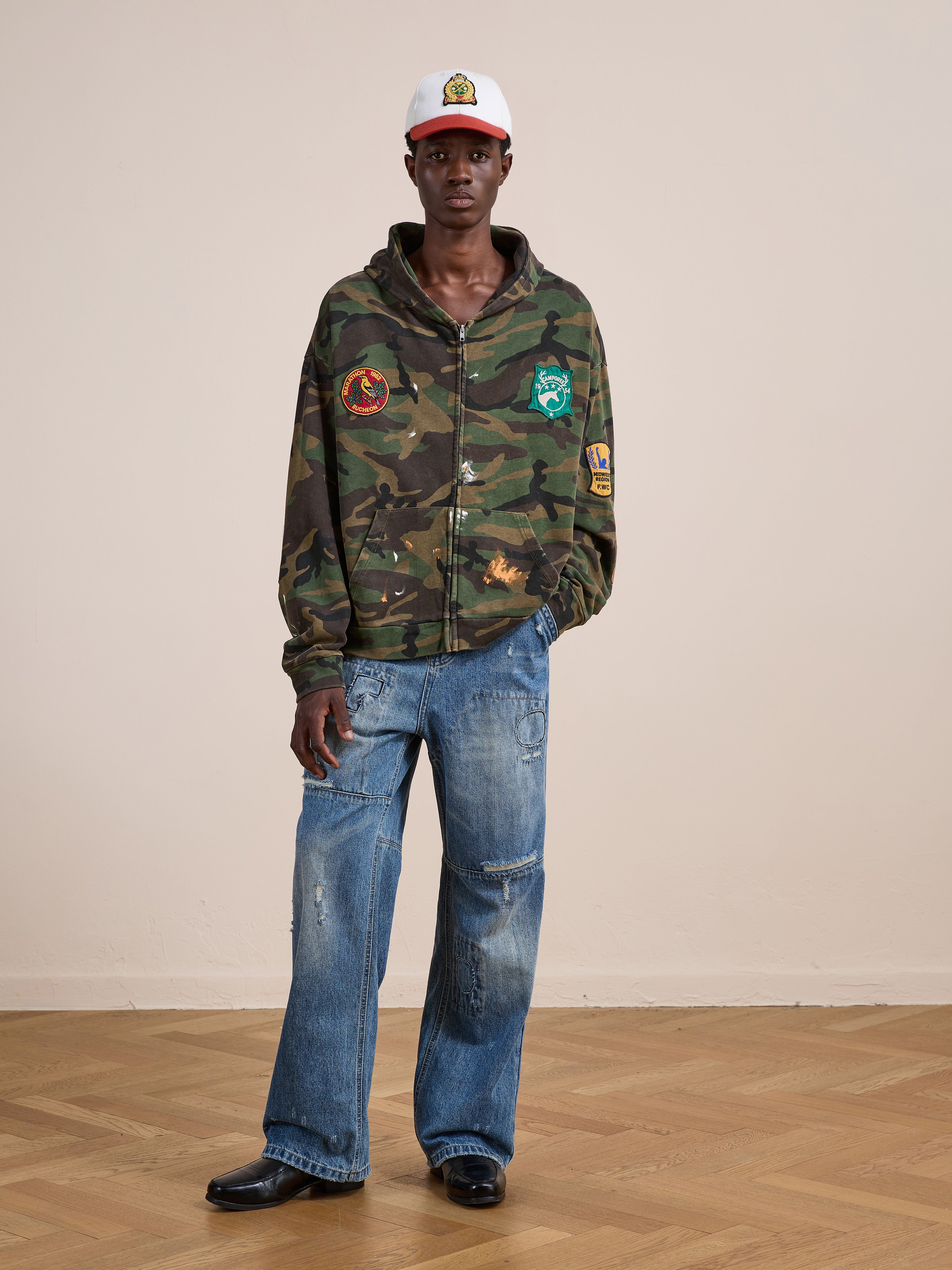 Indoors on a wooden floor, a person wears the FOUND Patch Camo Zip Up Hoodie in a relaxed fit, paired with loose blue jeans, black shoes, and a white baseball cap.