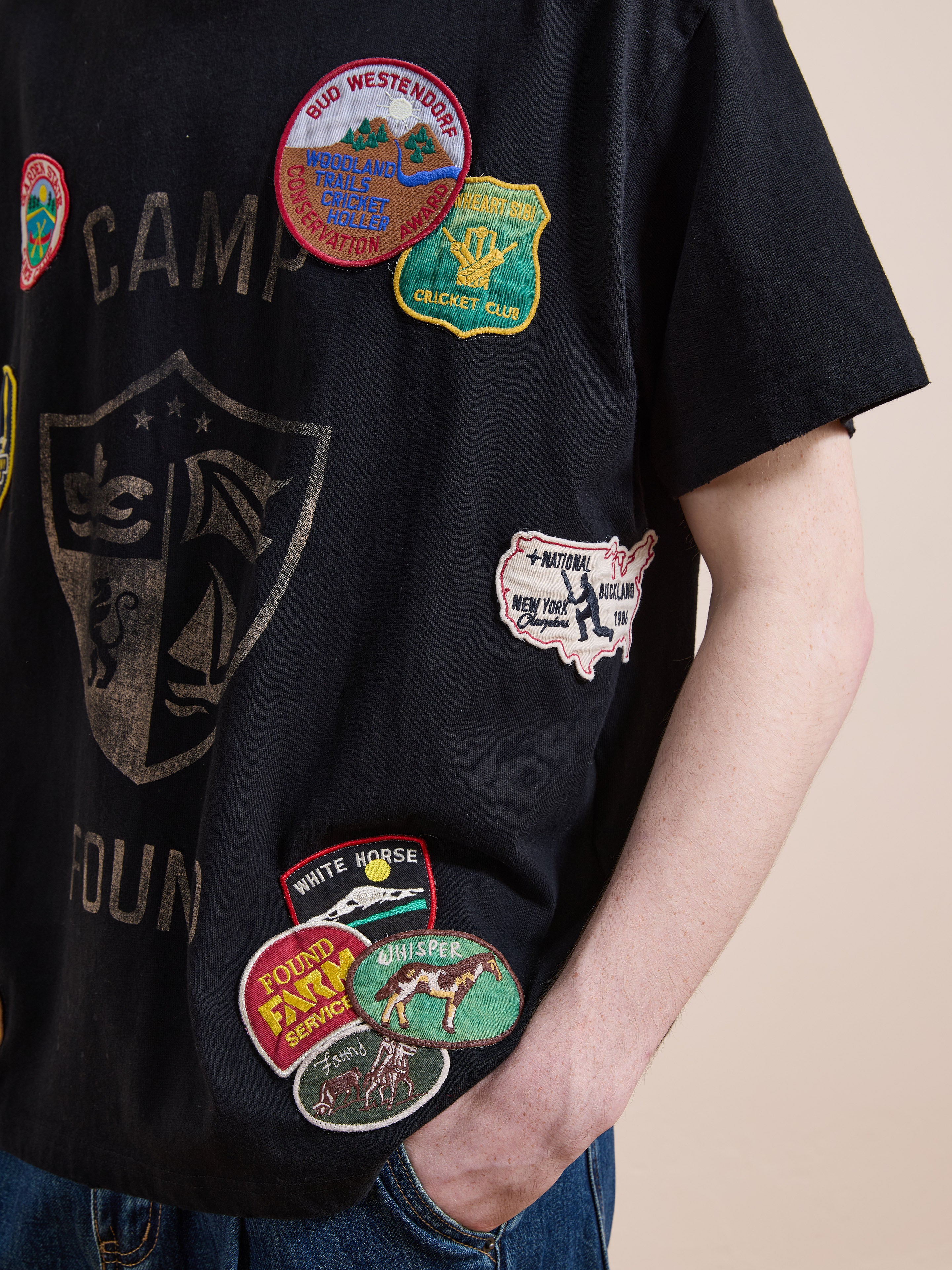 A person wears the FOUND Patchwork Camp Tee, a vintage black shirt with colorful embroidered patches, paired with blue jeans and one hand in their pocket.