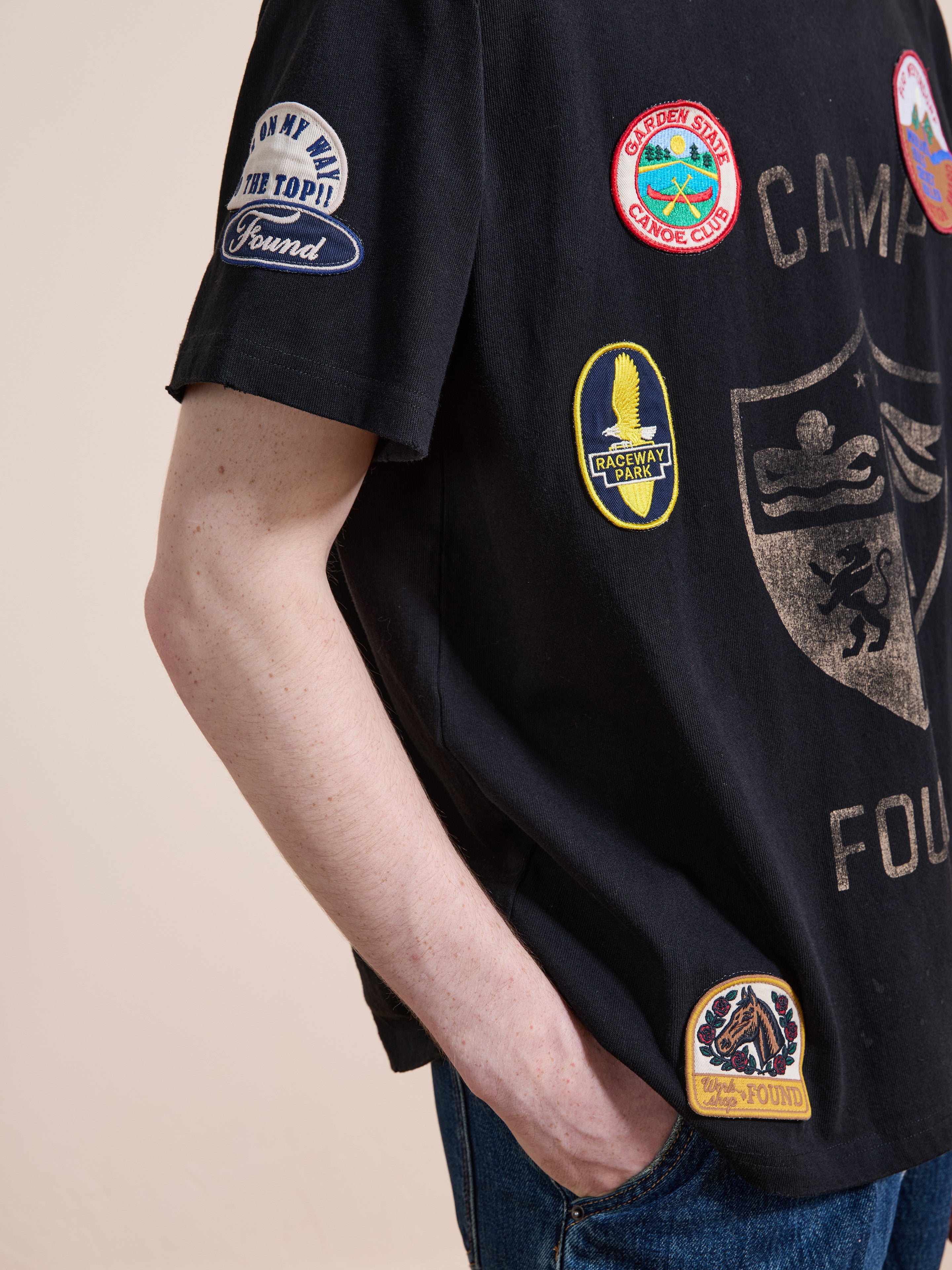 A person wearing the FOUND Patchwork Camp Tee, a vintage black shirt adorned with colorful patches, stands with one hand in their jean pocket.