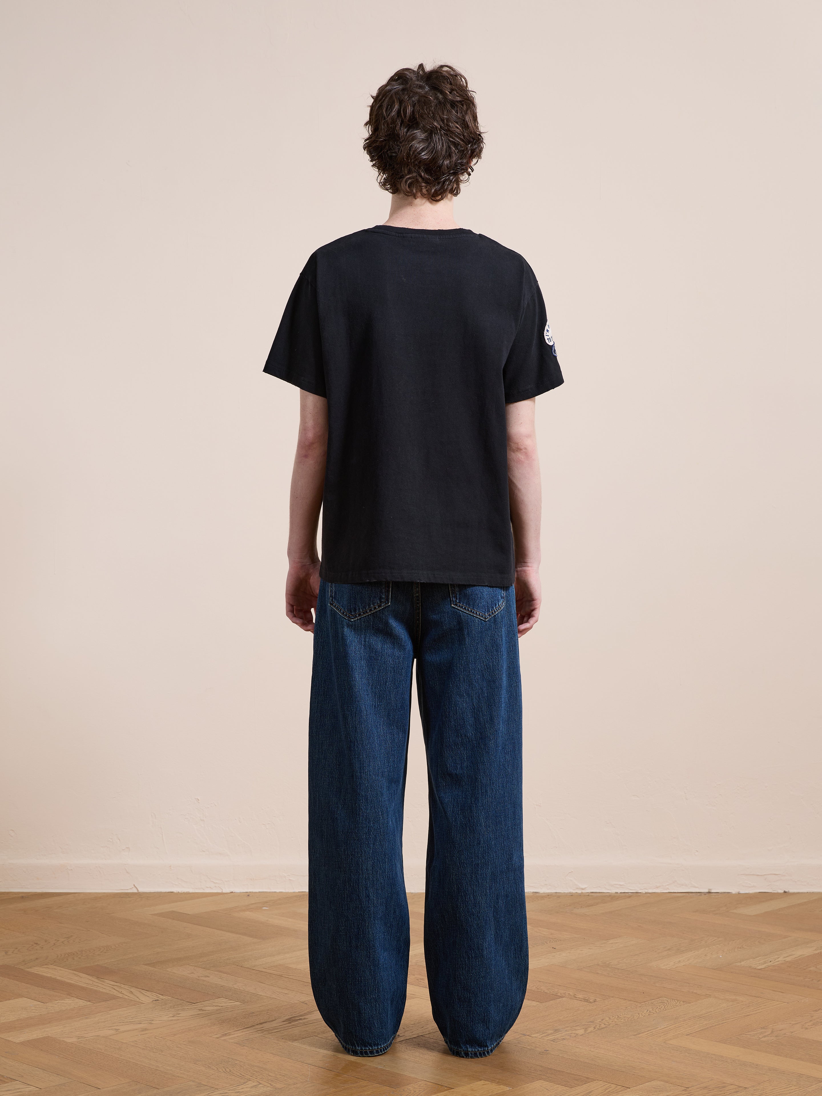 A person stands indoors on a wooden floor, facing away. They wear the FOUND Patchwork Camp Tee and loose-fitting blue jeans with a unisex relaxed fit, in front of a plain beige wall.