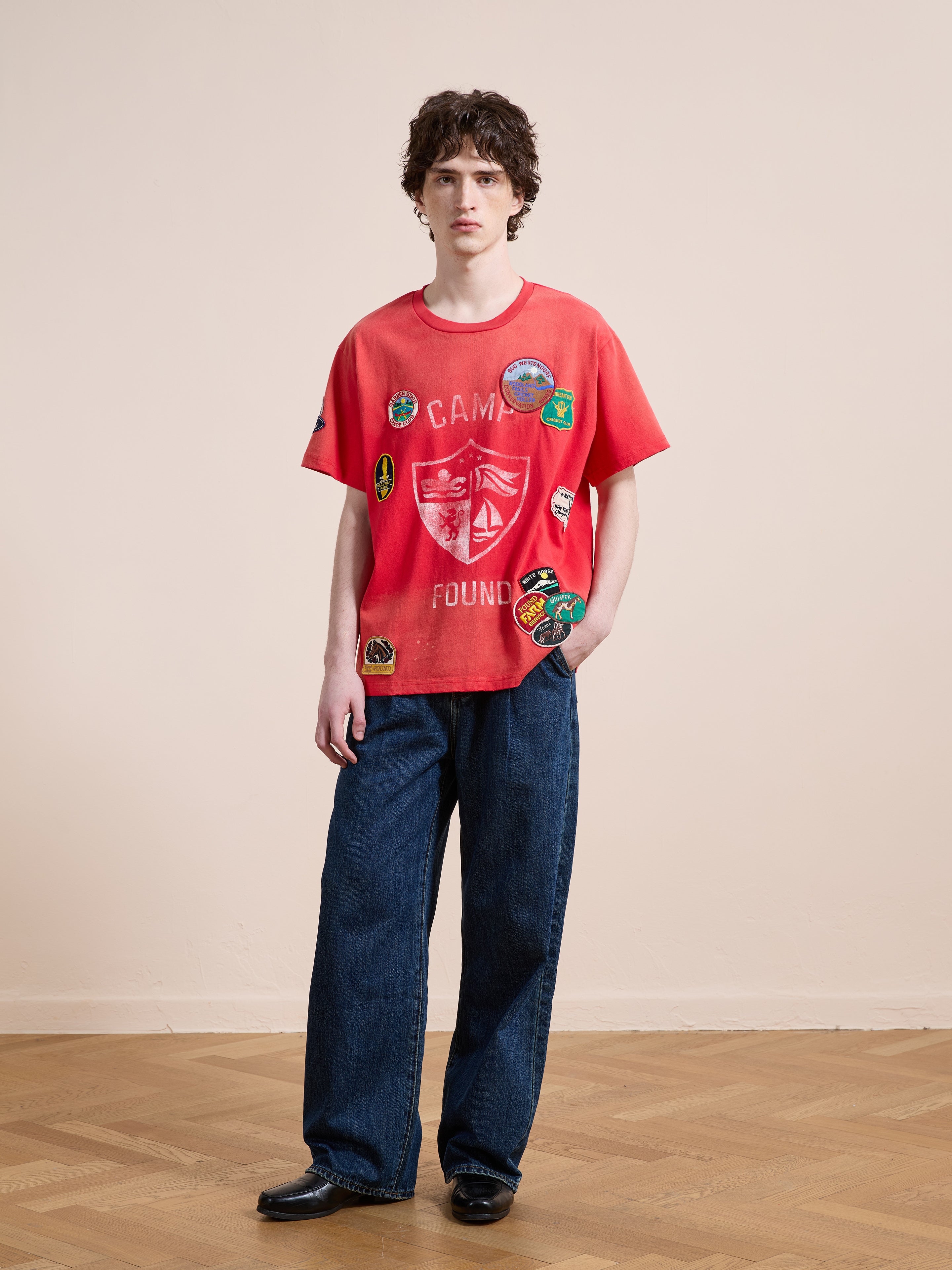 A person stands indoors against a plain background, wearing a relaxed fit FOUND Patchwork Camp Tee in red, paired with loose blue jeans and black shoes, looking directly at the camera.