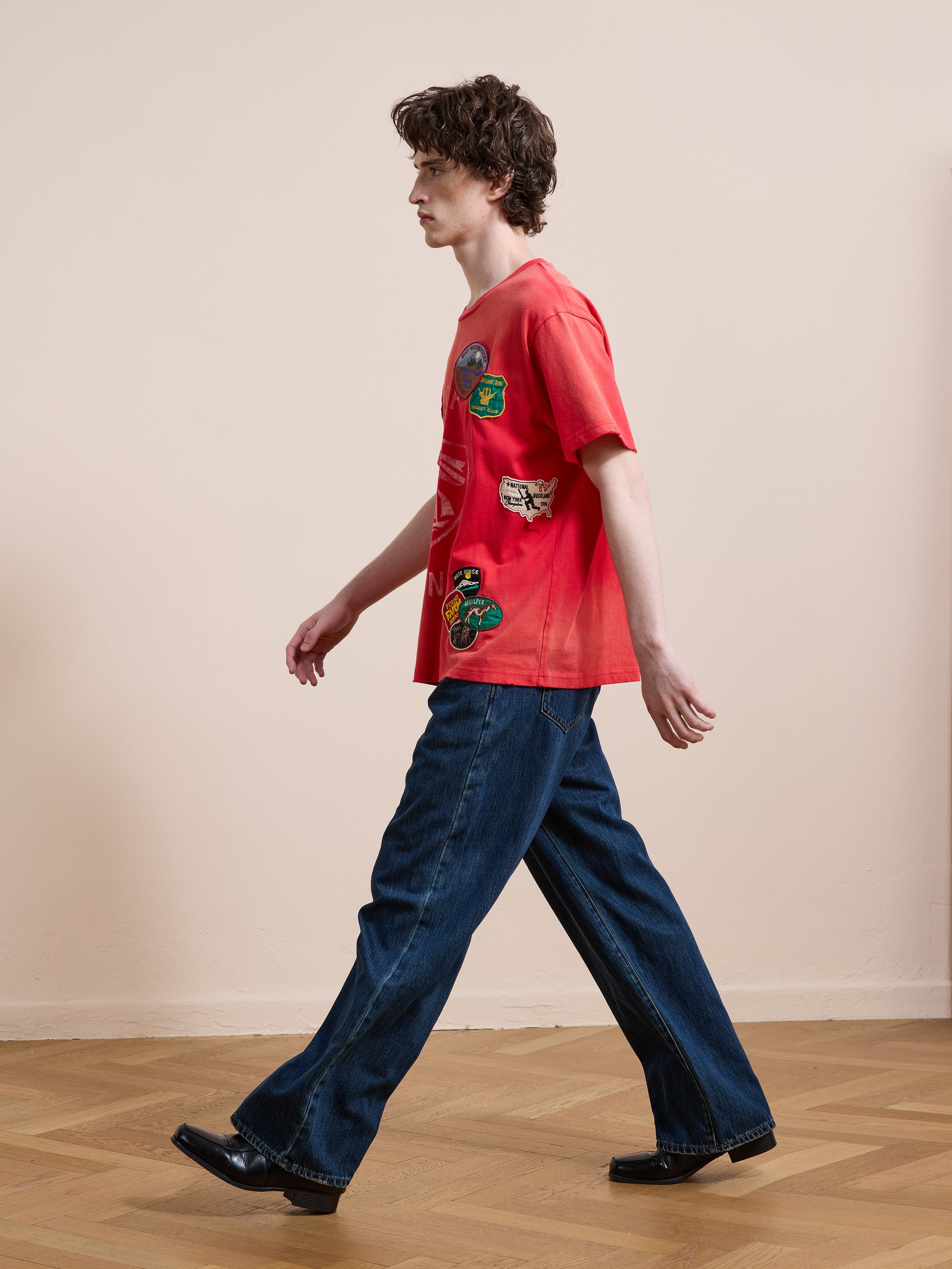 Indoors on a wooden floor, someone wears a relaxed fit FOUND Patchwork Camp Tee in washed red cotton, paired with wide-leg blue jeans and black shoes.