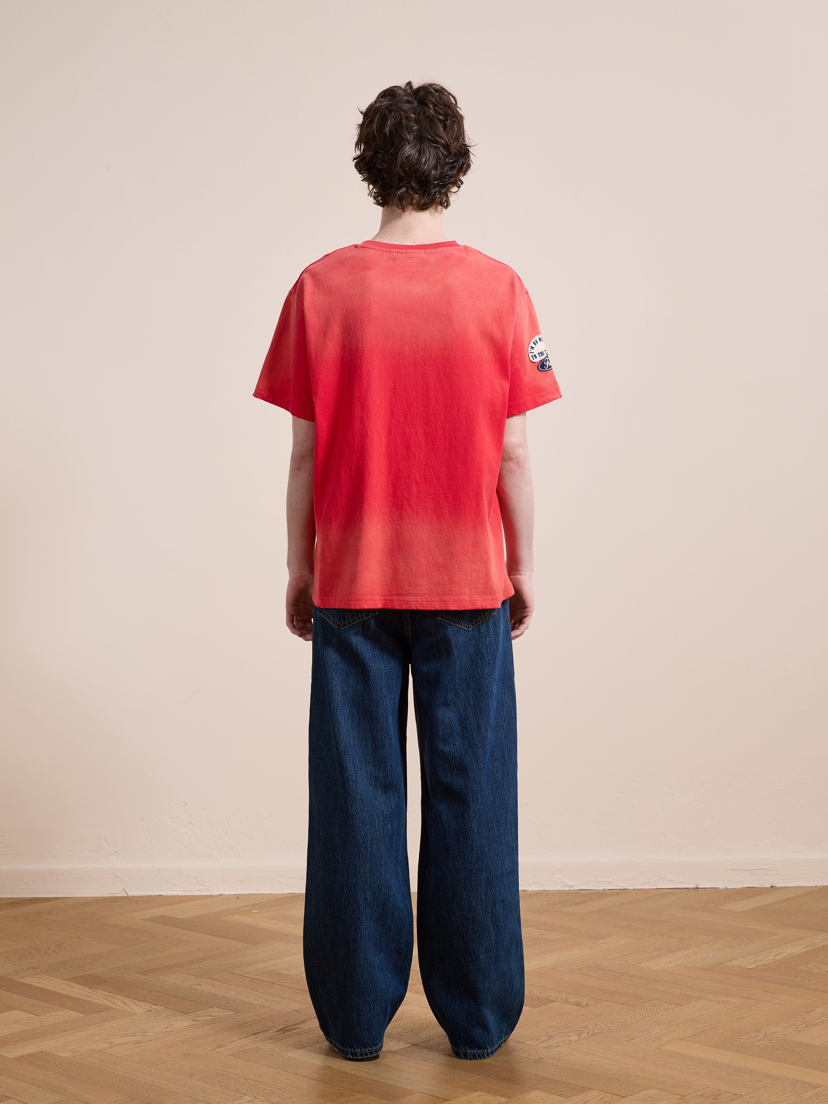 A person stands indoors with their back to the camera, wearing a FOUND Patchwork Camp Tee in washed red cotton and unisex relaxed fit, paired with wide-leg blue jeans on a parquet floor.