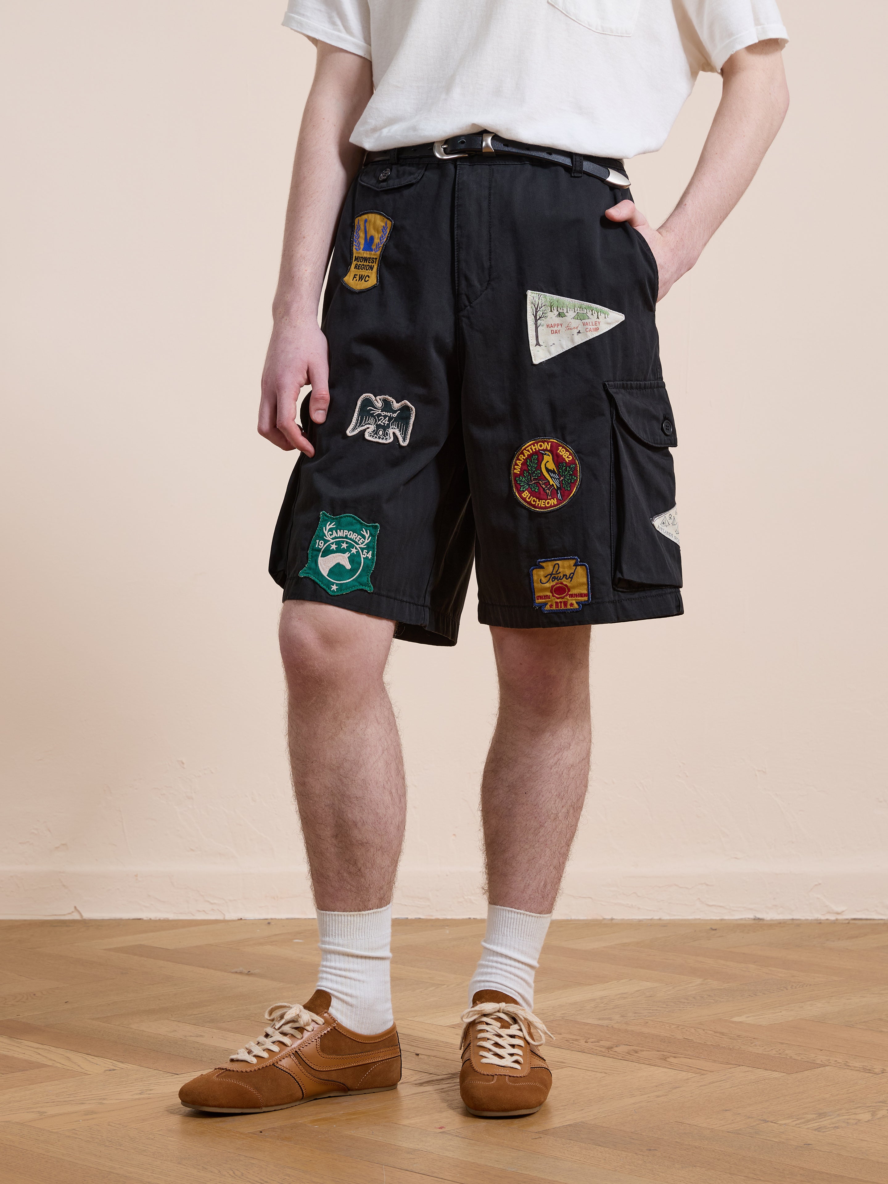 Someone stands on a wooden floor with one hand in their pocket, wearing a white t-shirt, Found Patchwork Cargo Shorts, white socks, and brown sneakers.