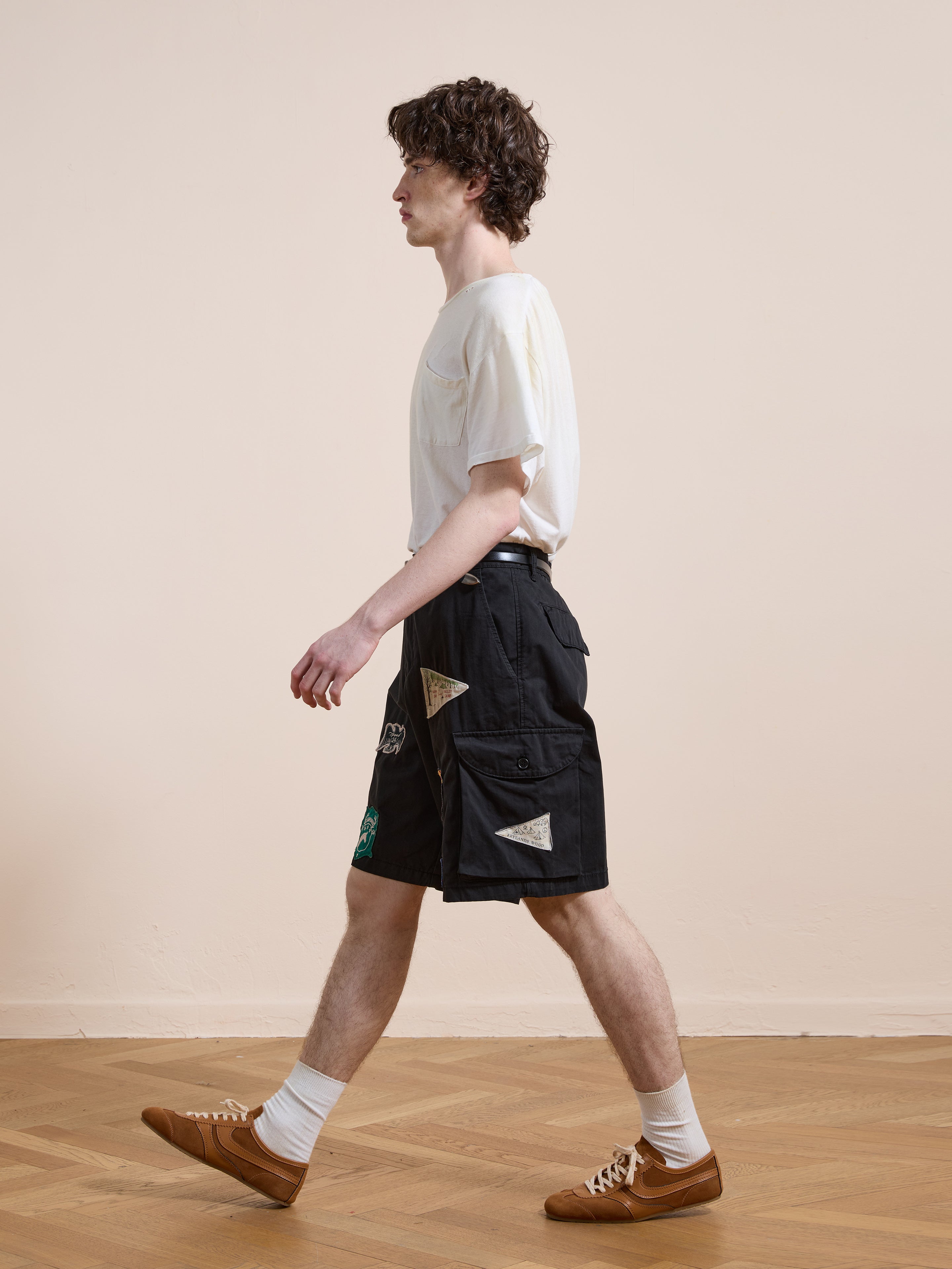 A man in a white t-shirt, Found Patchwork Cargo Shorts, white socks, and brown sneakers walks indoors on a wooden floor against a plain beige wall.