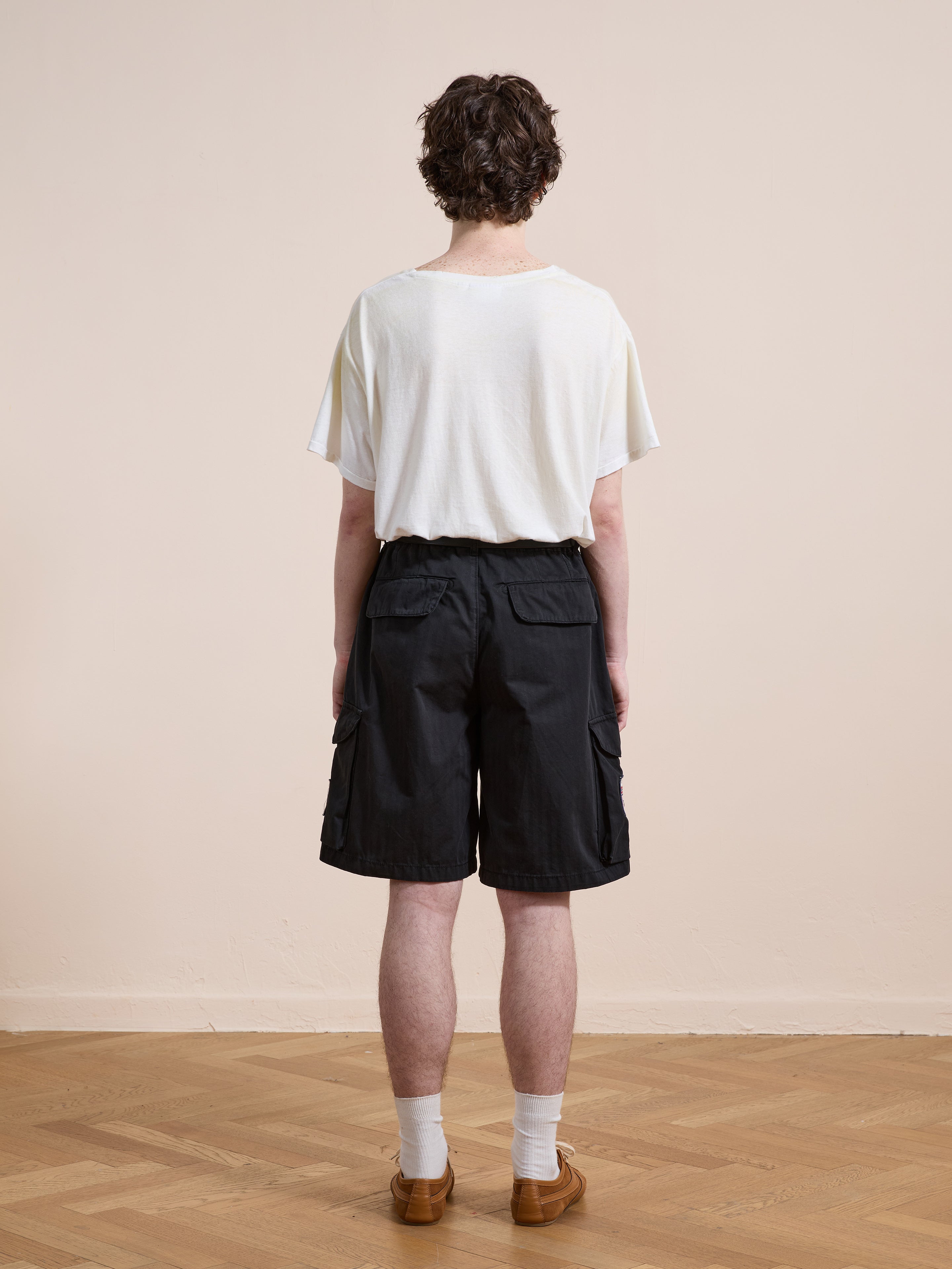 A person stands indoors on a wooden floor, facing away, wearing Found Patchwork Cargo Shorts with a white T-shirt, white socks, and brown shoes.