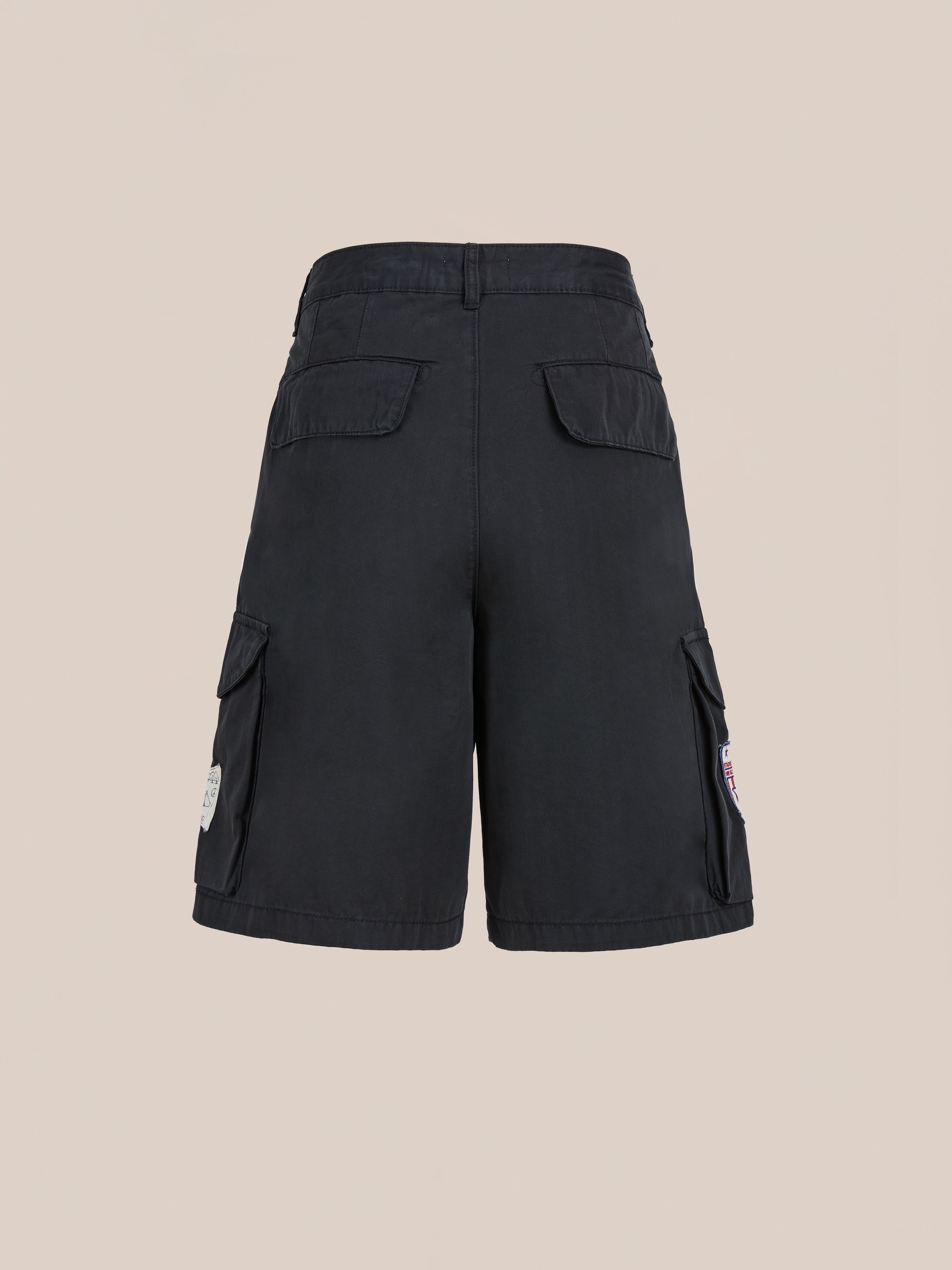 The Found Patchwork Cargo Shorts are shown from the back in vintage black with flap and side cargo pockets featuring patches, displayed on a beige background. Unisex relaxed fit offers versatile styling.