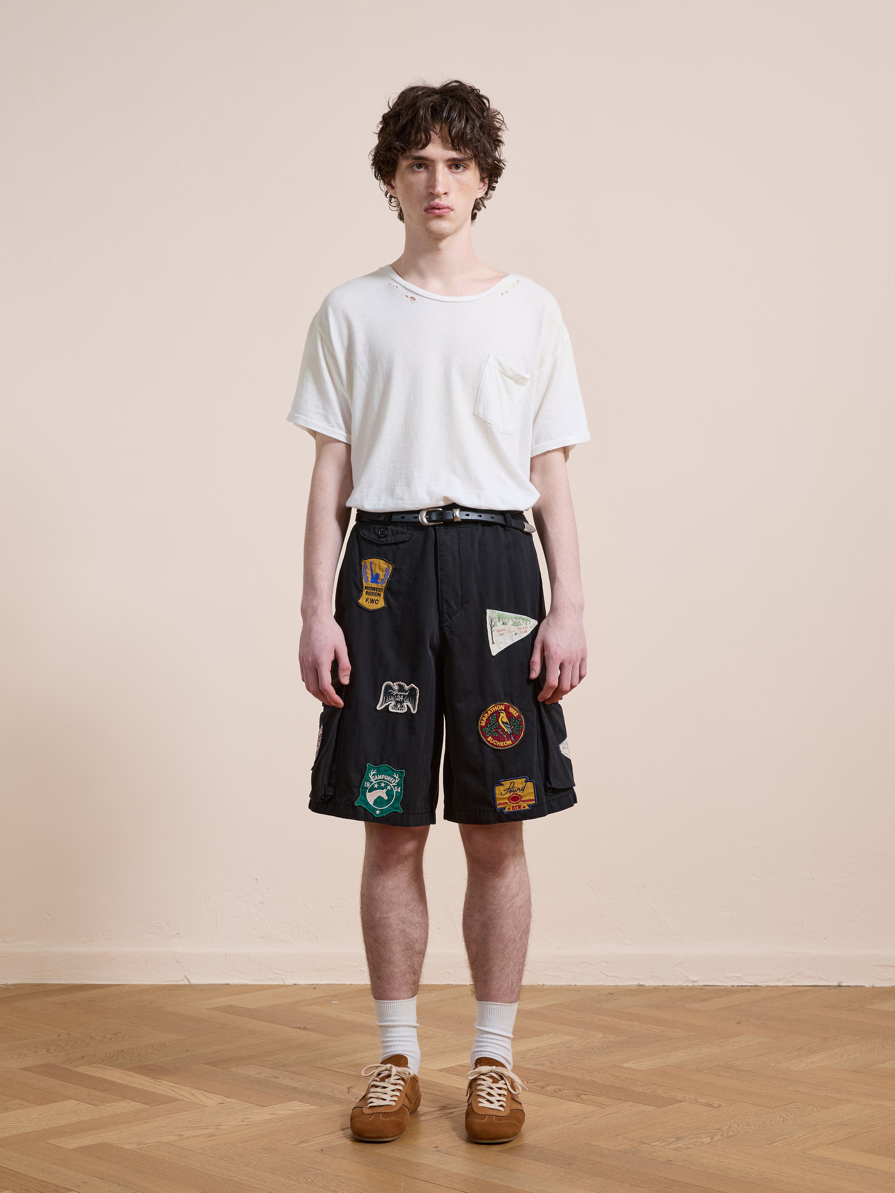 A young person stands indoors facing forward against a plain background, wearing Found's Patchwork Cargo Shorts in a vintage black wash with a relaxed unisex fit, paired with a white t-shirt, white socks, and brown shoes.