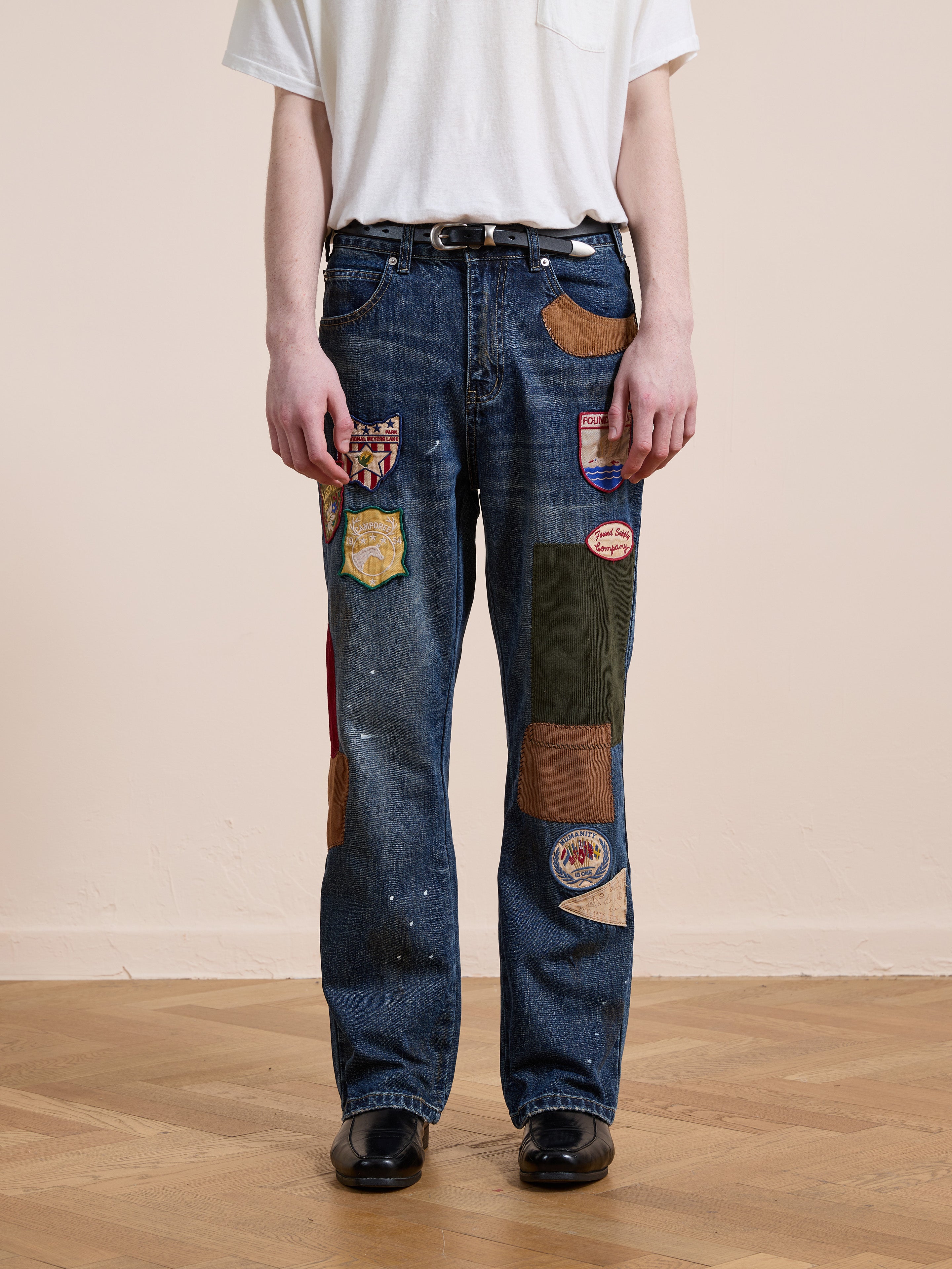 A person in a white t-shirt, FOUND Patchwork Jeans, a black belt, and black shoes stands on a wooden floor against a neutral wall.