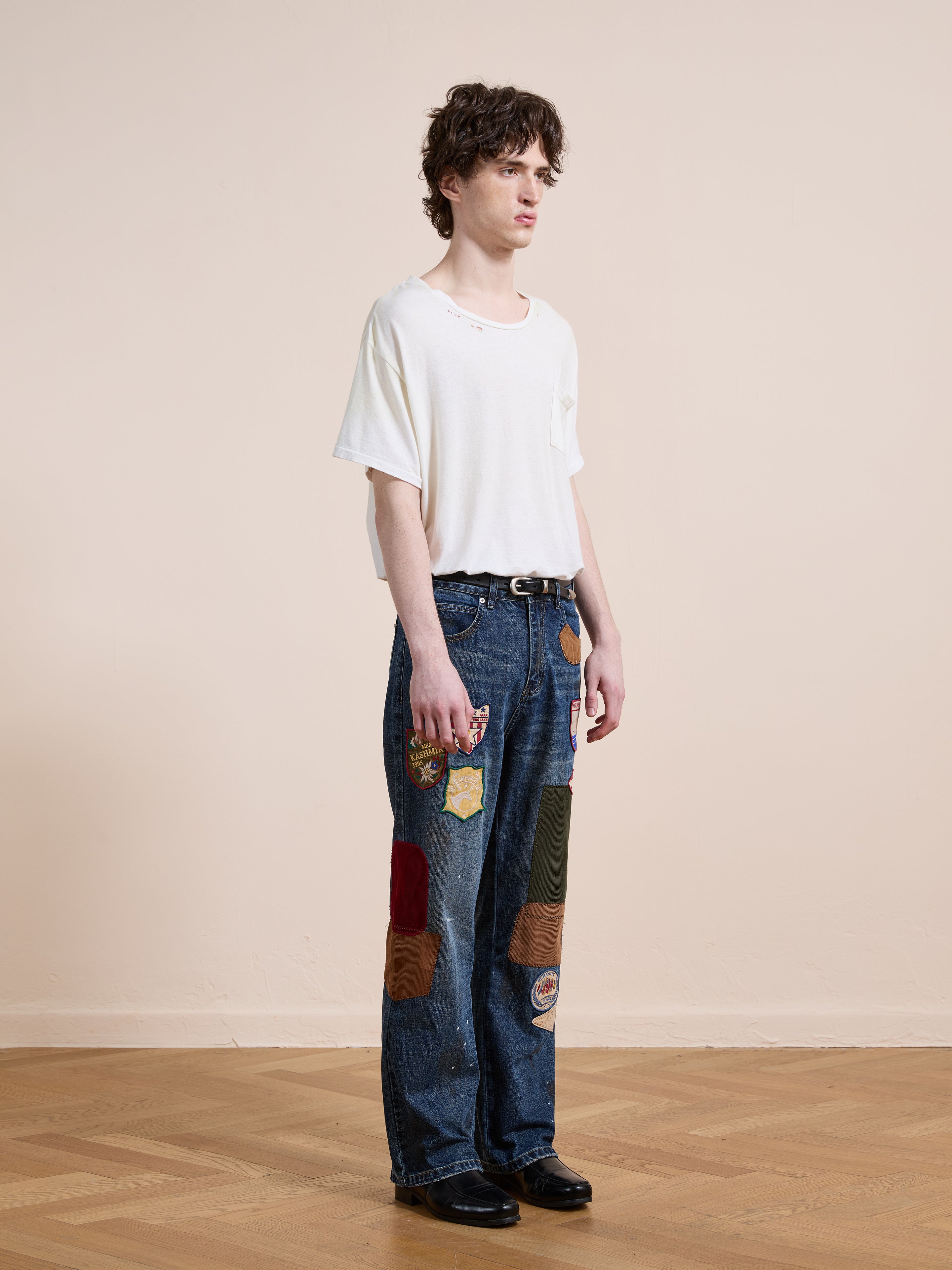 A young man stands indoors wearing FOUND Patchwork Jeans—blue pleated, relaxed fit with colorful fabric patches—paired with a loose white T-shirt and black shoes.
