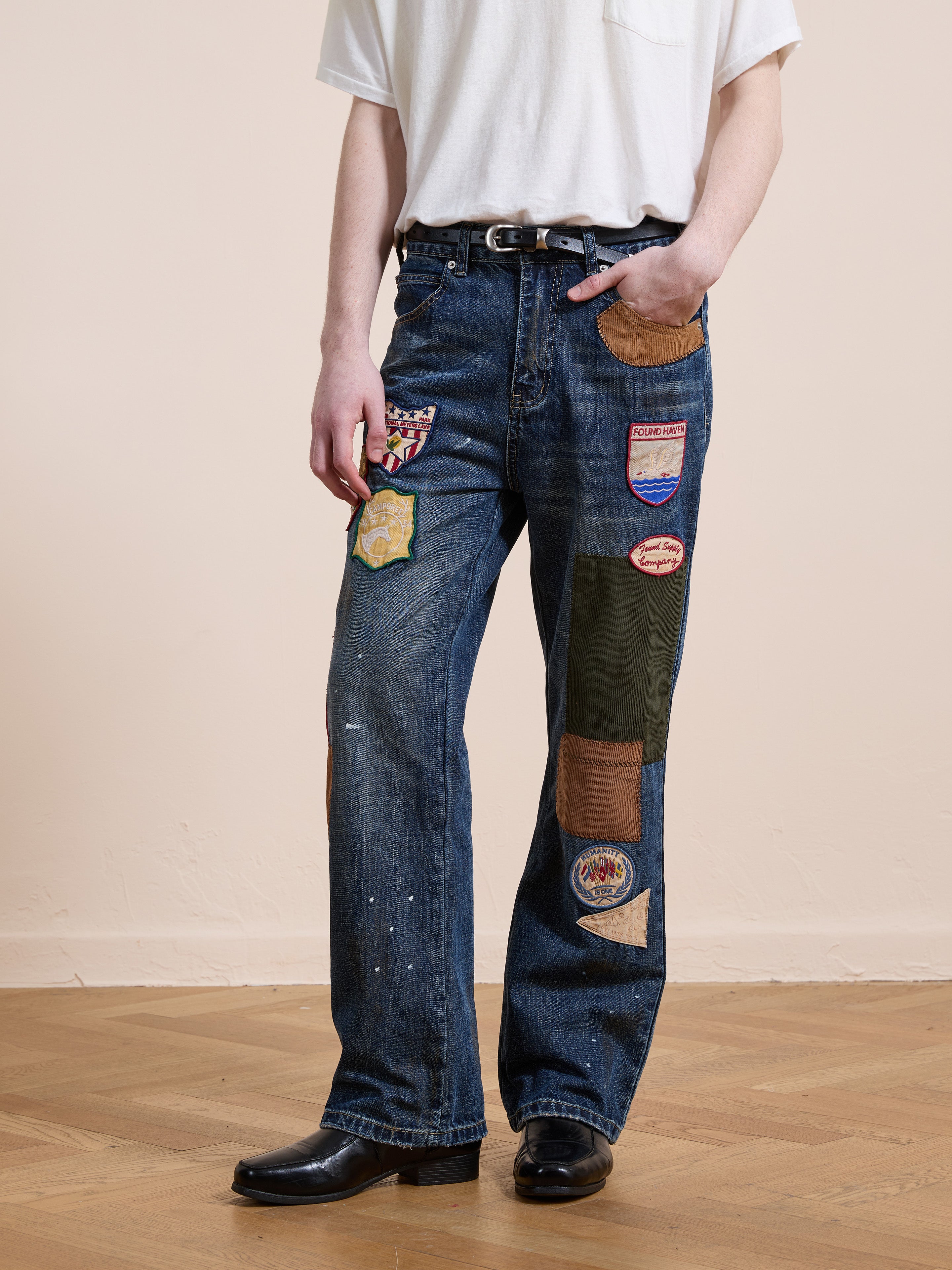A person stands on a wooden floor with one hand in their pocket, wearing a white T-shirt, black belt, black shoes, and FOUND’s Patchwork Jeans.