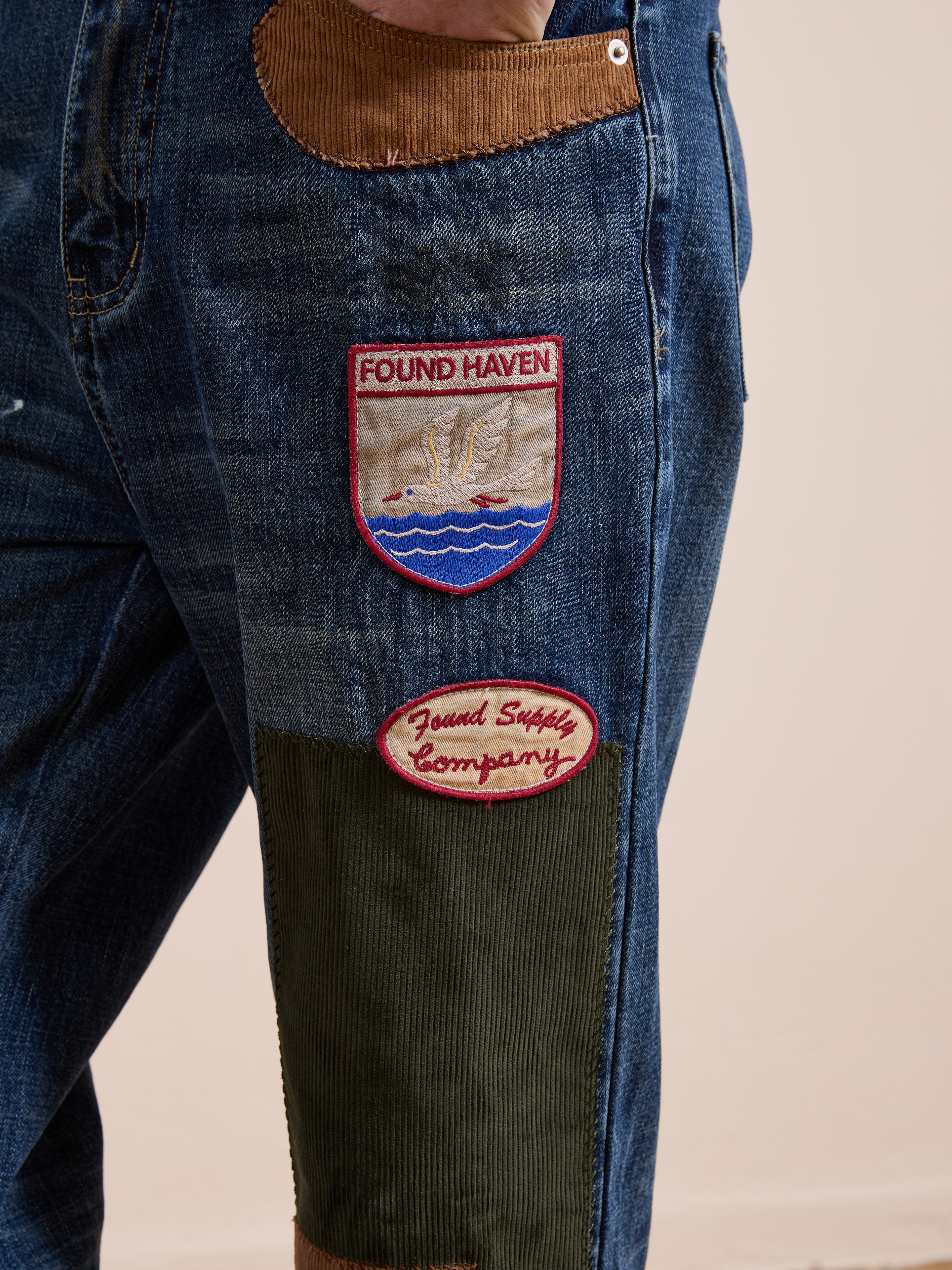 Close-up of FOUND Patchwork Jeans, vintage denim with two embroidered patches: one says "FOUND HAVEN" with a graphic, the other "Found Supply Company." Green and brown fabric accents add flair to these relaxed fit jeans.