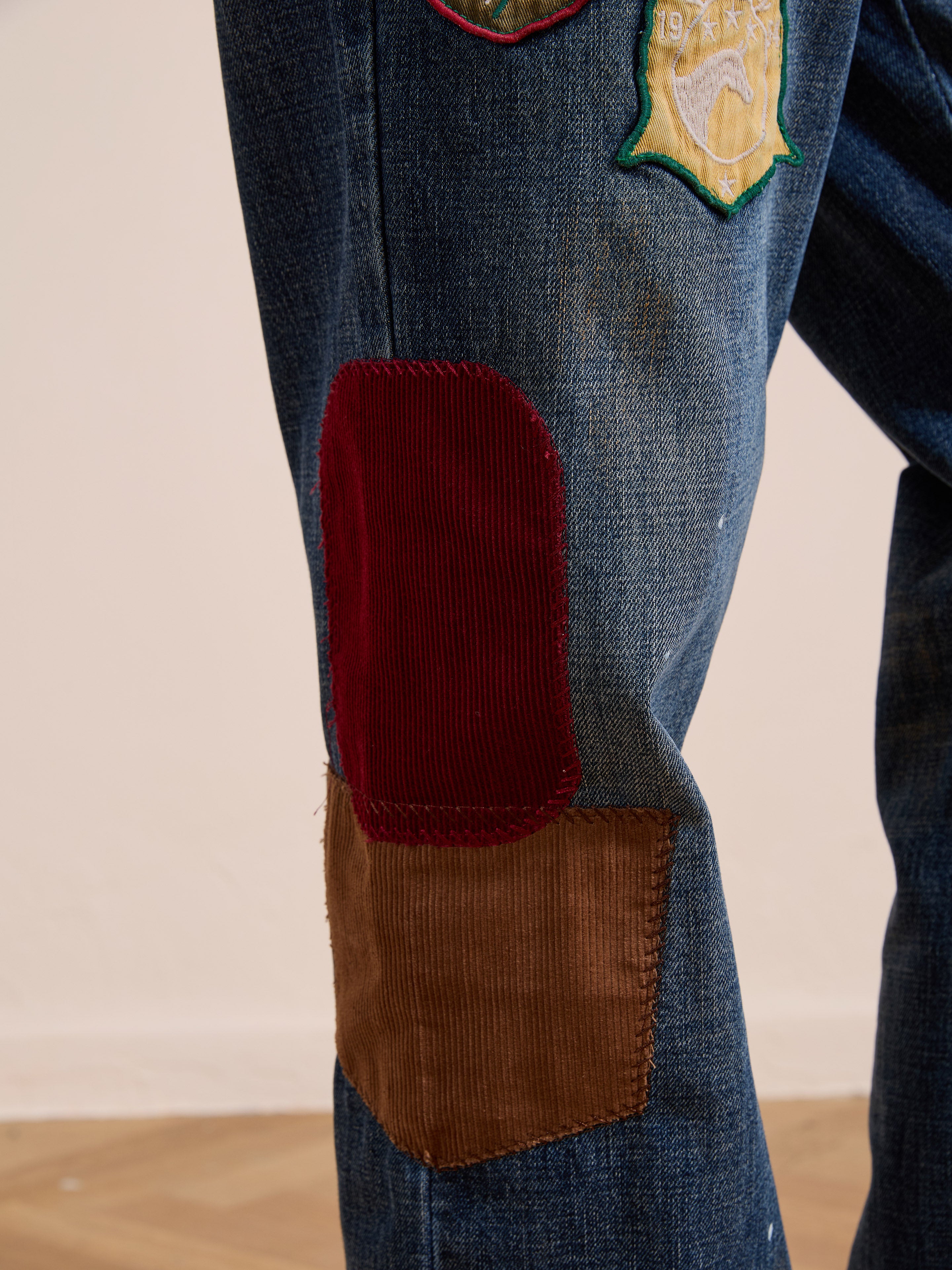 Close-up of FOUND Patchwork Jeans, crafted from vintage denim in a relaxed fit, with red corduroy and brown patches on the knee and a badge patch above.