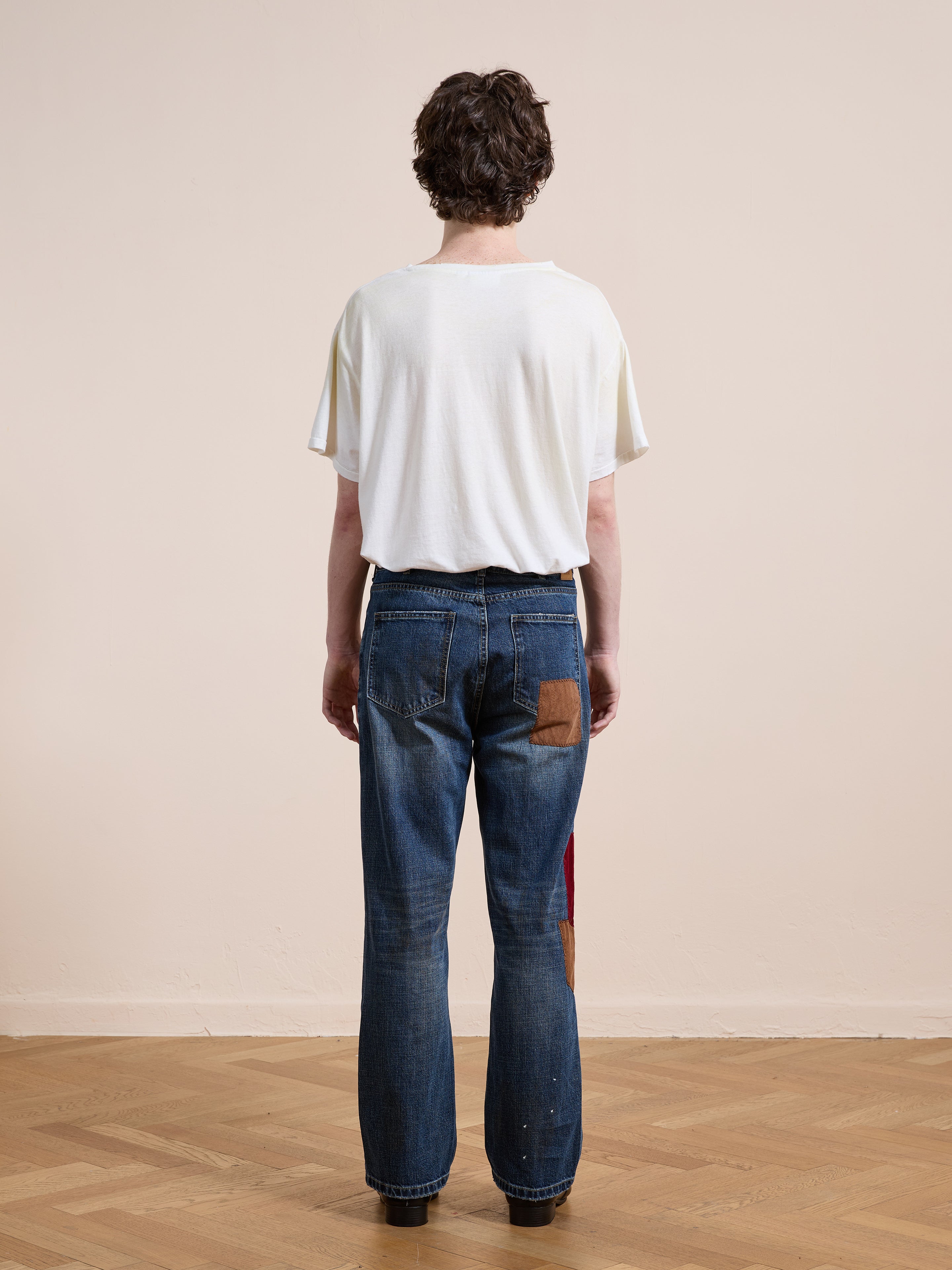 A person stands indoors on a wooden floor, facing away from the camera, wearing a white t-shirt and FOUND Patchwork Jeans featuring vintage-inspired relaxed fit and visible patches.