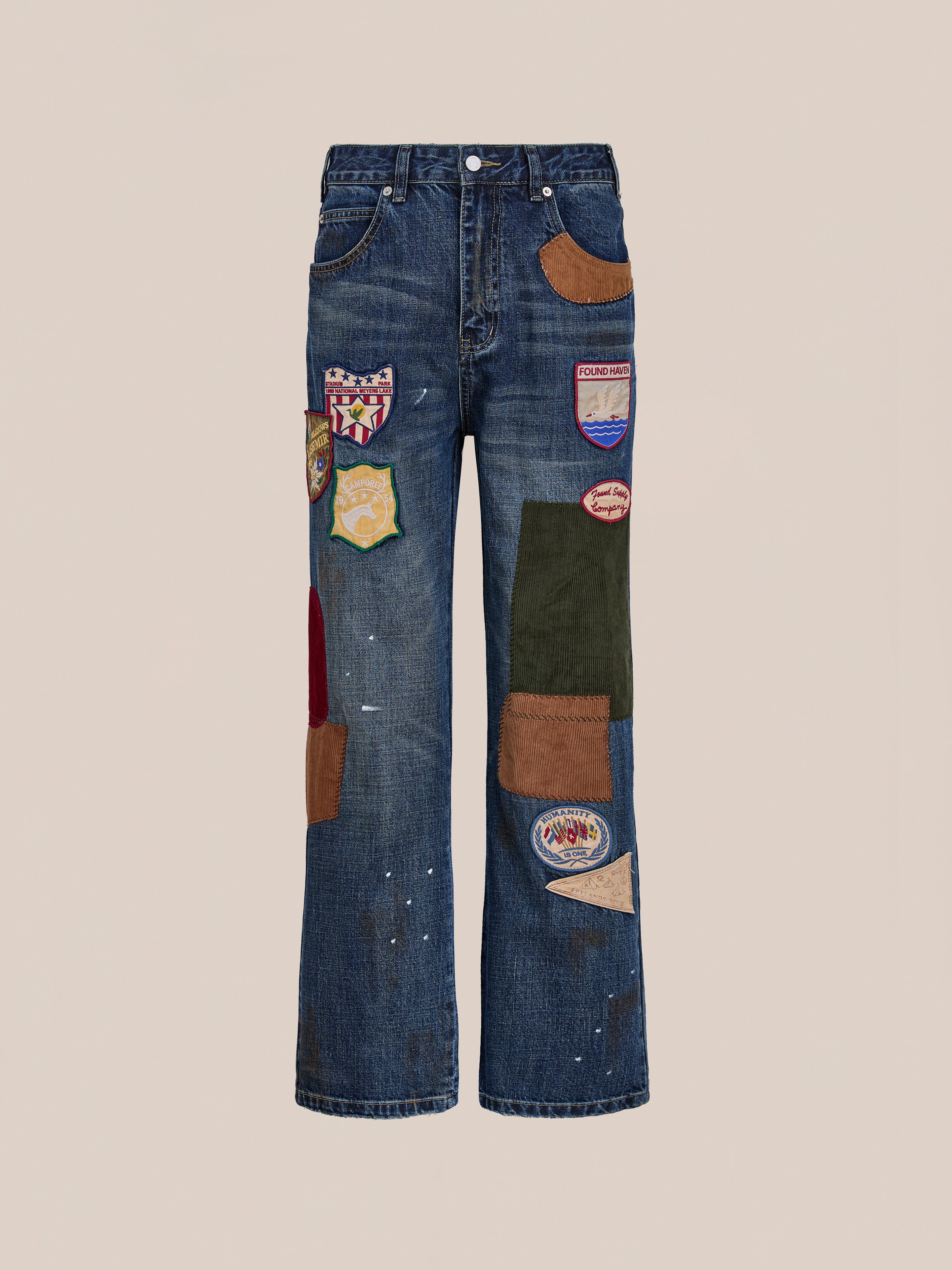The FOUND Patchwork Jeans feature colorful fabric patches and badges sewn on the front, creating a unique vintage look. Displayed against a plain beige background.