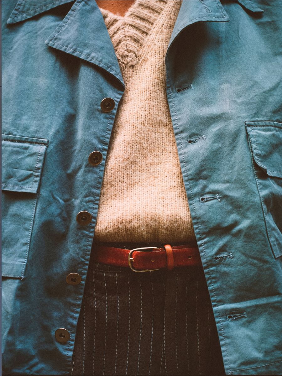 The individual radiates vintage workwear charm in a Found Patina Work Jacket over a beige knit sweater, paired with pinstriped pants and a brown leather belt.