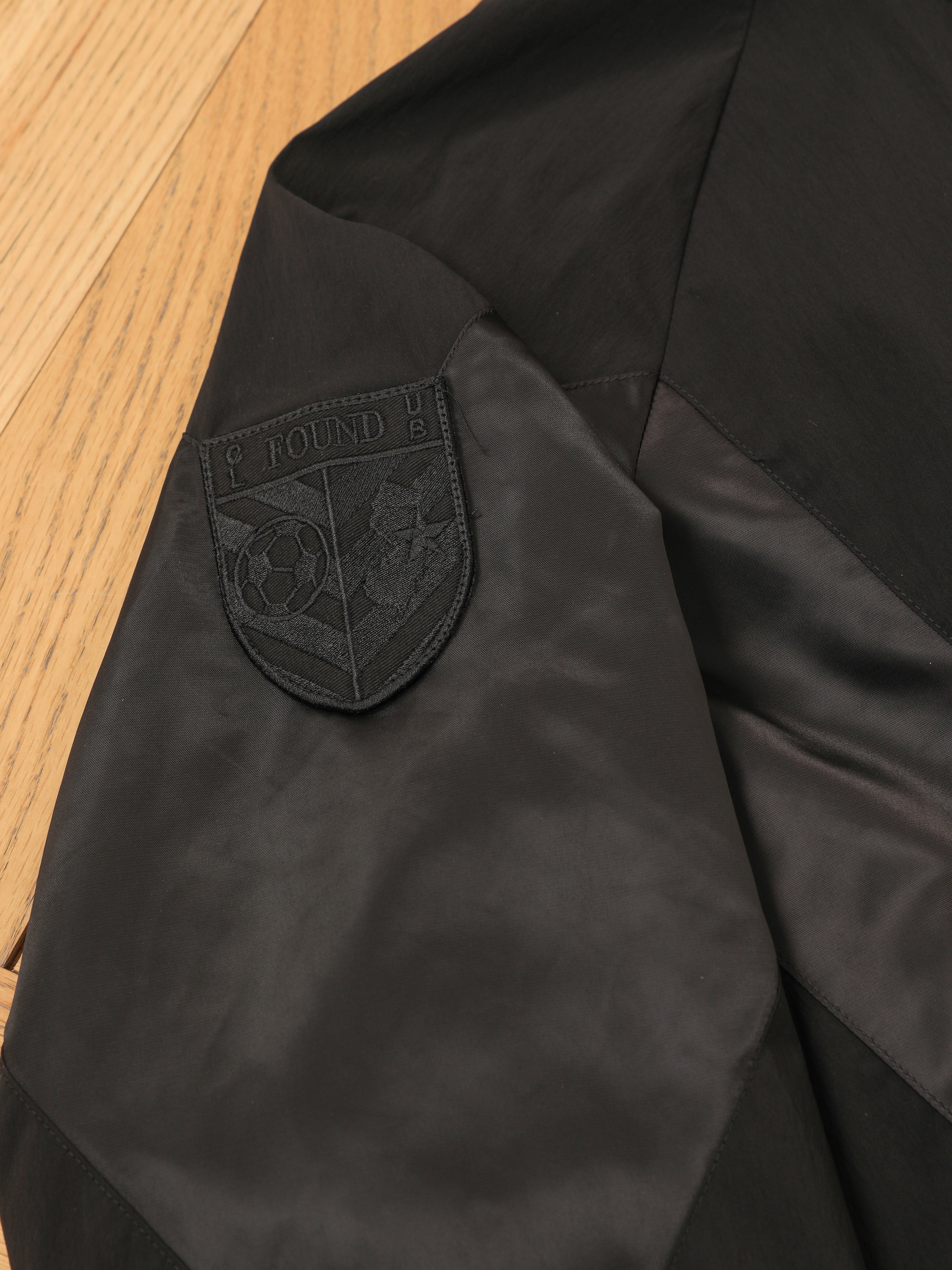 The Pine and Rose Jacket by FOUND features a black patch with a soccer emblem and "FOUND" text, sewn onto the sleeve of this black vintage sportswear track jacket, shown on a wooden surface.