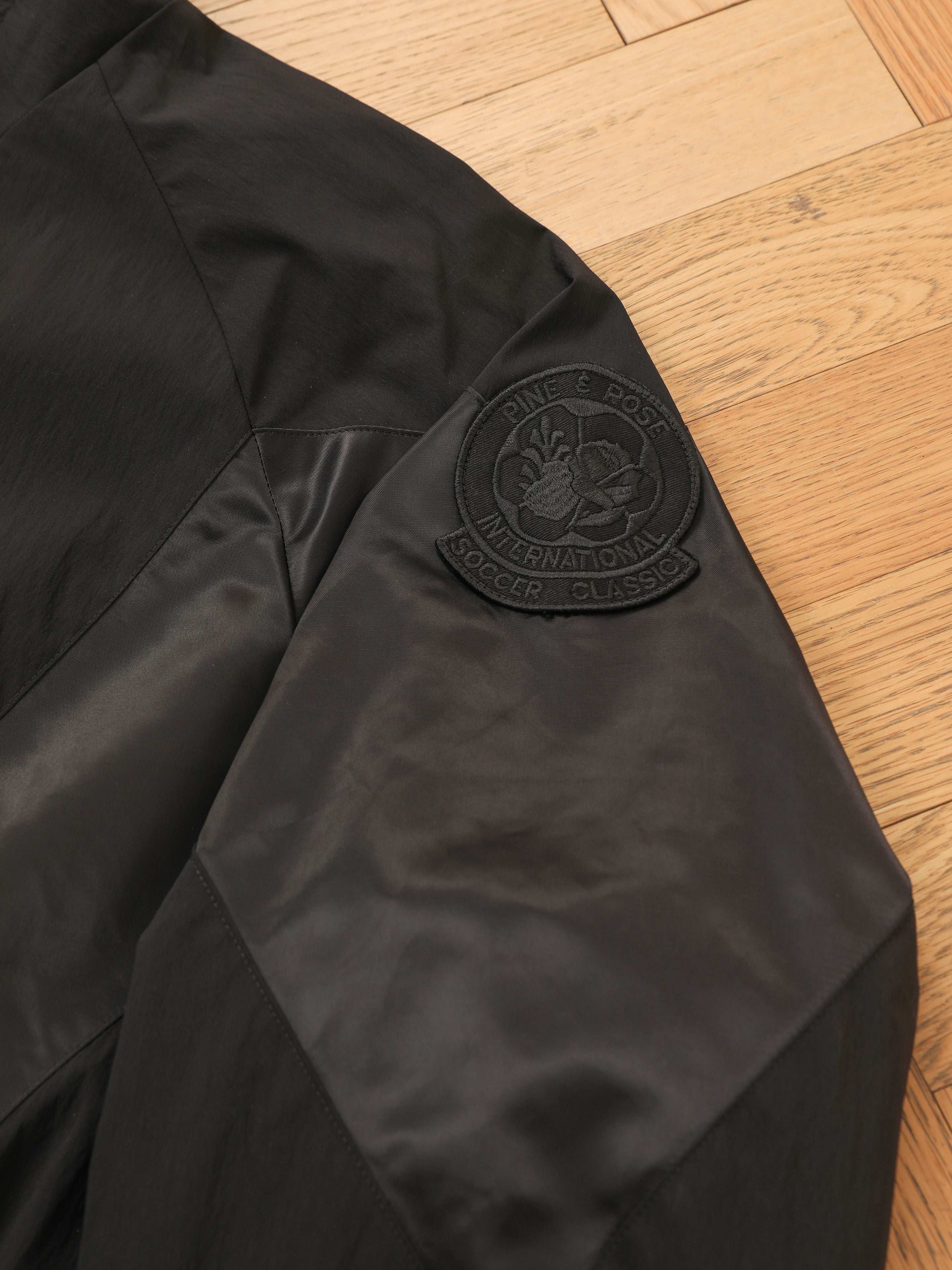 A close-up of the FOUND Pine and Rose Jacket, a vintage sportswear black track jacket, featuring a round embroidered “Pine & Post International Soccer Classic” patch on the sleeve, laid on a wooden floor.