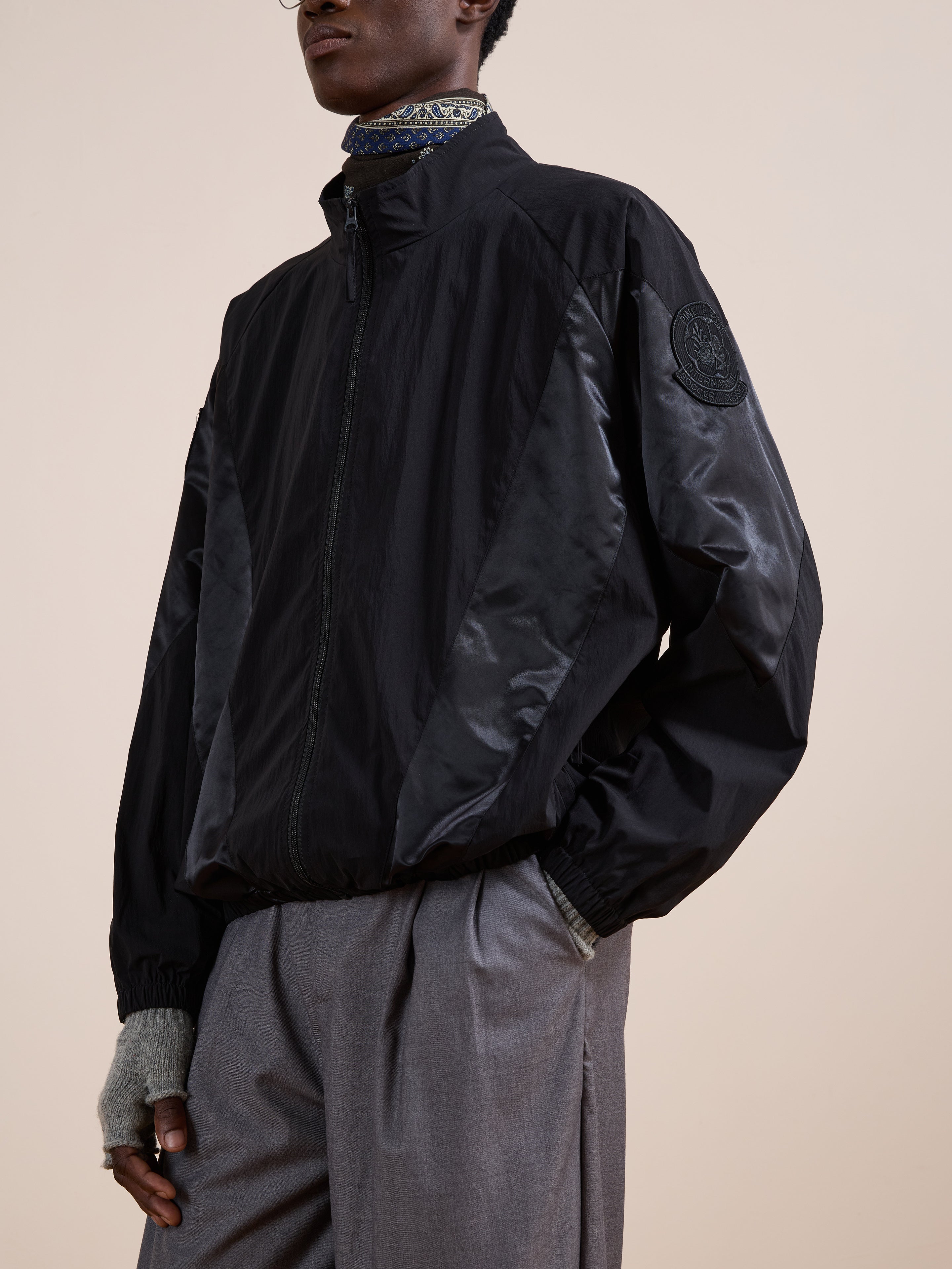 A person wears the FOUND Pine and Rose Jacket, a black unisex windbreaker with a sleeve patch, paired with grey wide-leg trousers and fingerless gloves. Only their lower face and torso are shown.